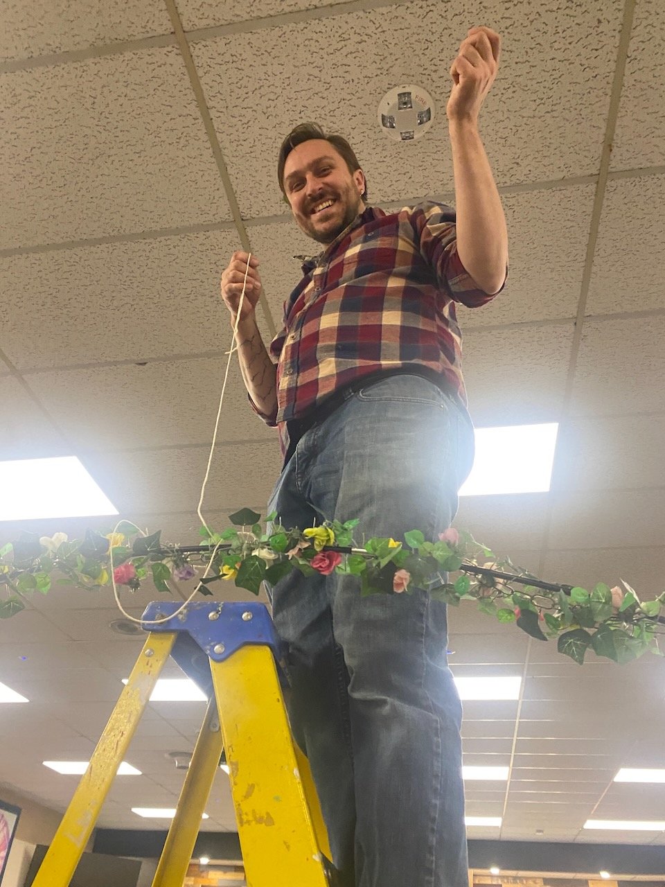 Forrest Hejkal, member at large of The Healing Feast Foundation board, is standing on a yellow ladder, smiling, holding a string, and surrounded by decorative flowers and greenery in an indoor setting with ceiling tiles and fluorescent lights.