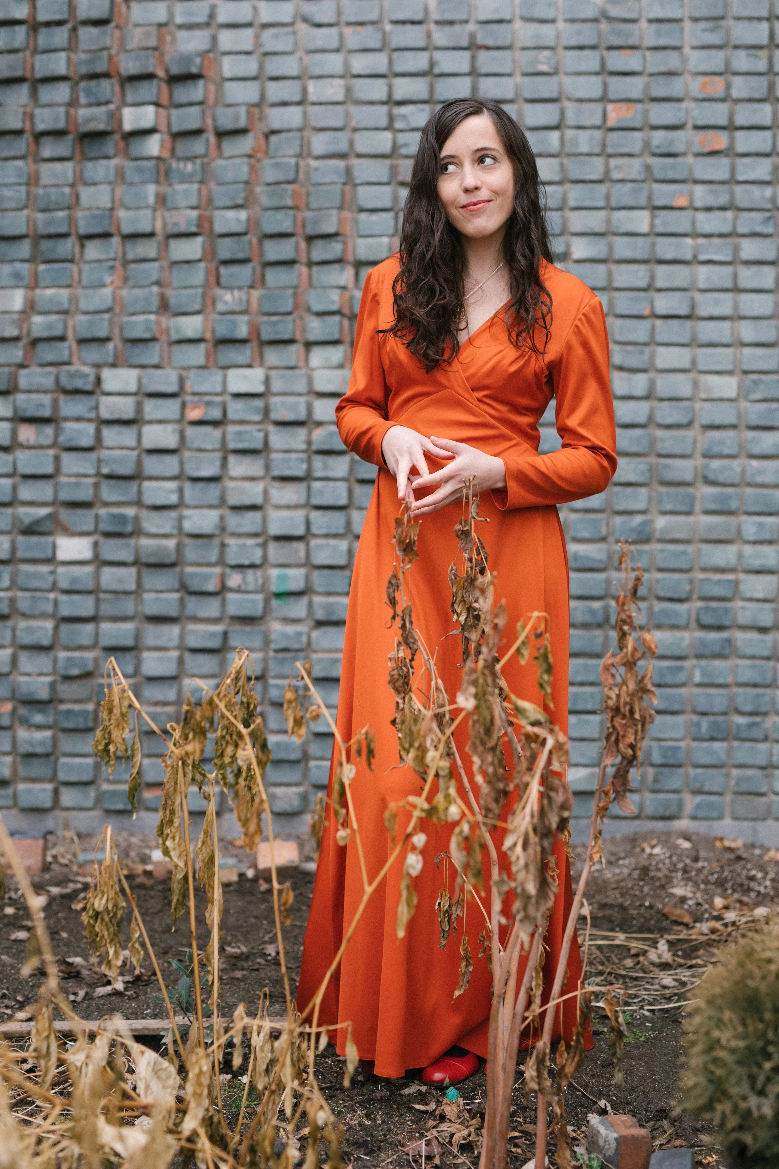 Sari Brown, founder and director of The Healing Feast, is a woman with long dark wavy hair wearing an orange dress standing in a garden with dried plants and a gray brick wall in the background.