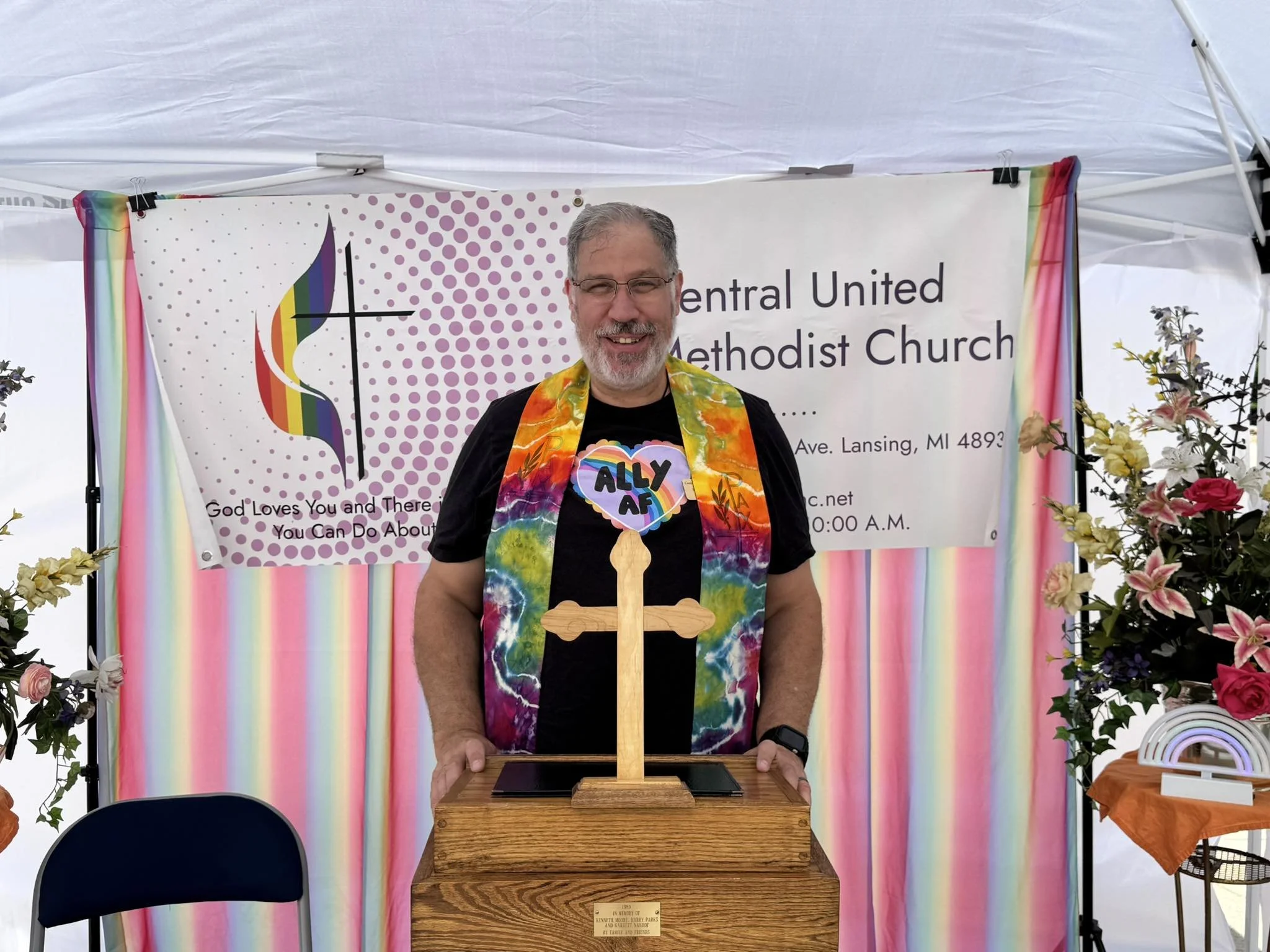 Rich Burstall, secretary of The Healing Feast Foundation board, a man with glasses and gray hair wearing a rainbow tie-dye stole, stands in front of a banner for the United Methodist Church with rainbow decorations, at an LGBTQ pride event.