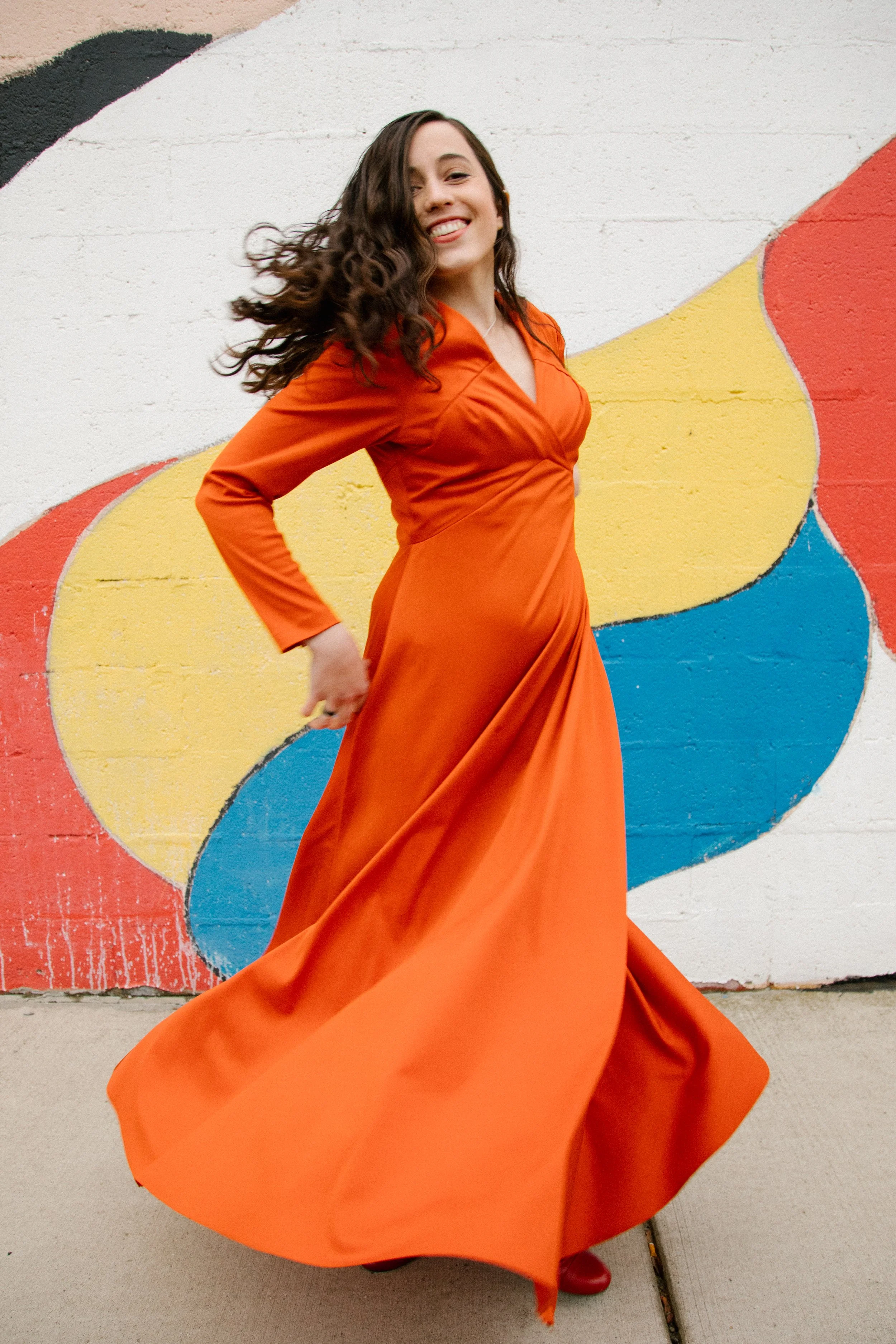 Sari Brown, founder and director of The Healing Feast, is a woman with long, curly brown hair wearing a flowing orange dress, standing in front of a colorful mural wall, twirling and smiling, looking at the camera.