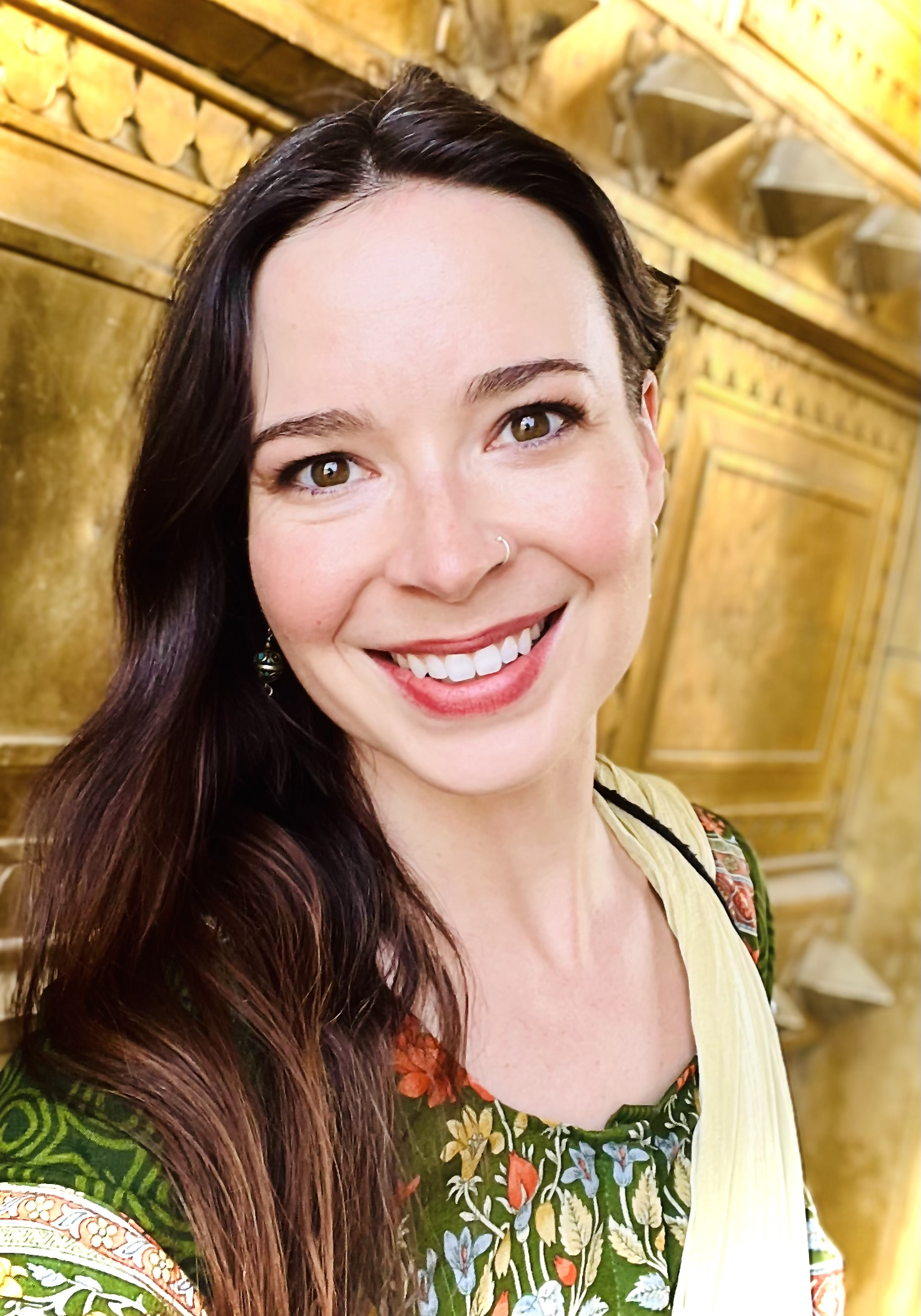 Katie Lynne, member at large of The Healing Feast Foundation board, is a smiling woman with long dark hair, wearing Indian clothing with floral patterns, standing in front of a wooden interior with intricate carvings.