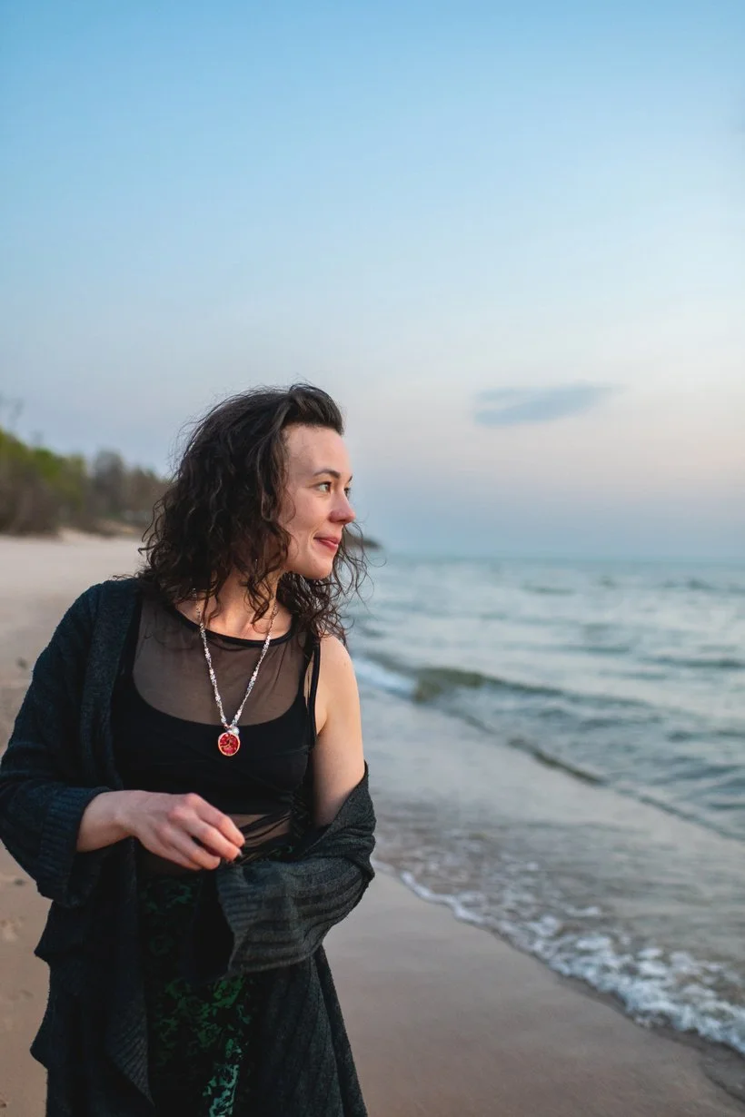 A woman with curly dark hair walking along the beach during sunset, wearing a black top with a sheer overlay and a pendant necklace.