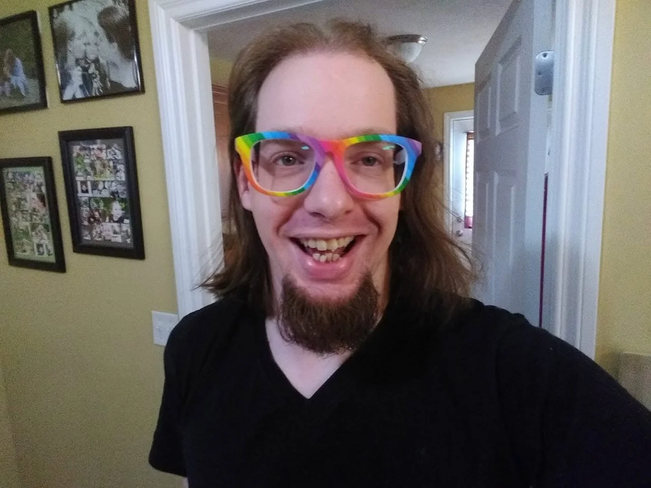 David LaLone, treasurer of The Healing Feast Foundation board, is a man with shoulder-length brown hair, a goatee, wearing rainbow-colored glasses, smiling in a home interior with framed pictures on a yellow wall.