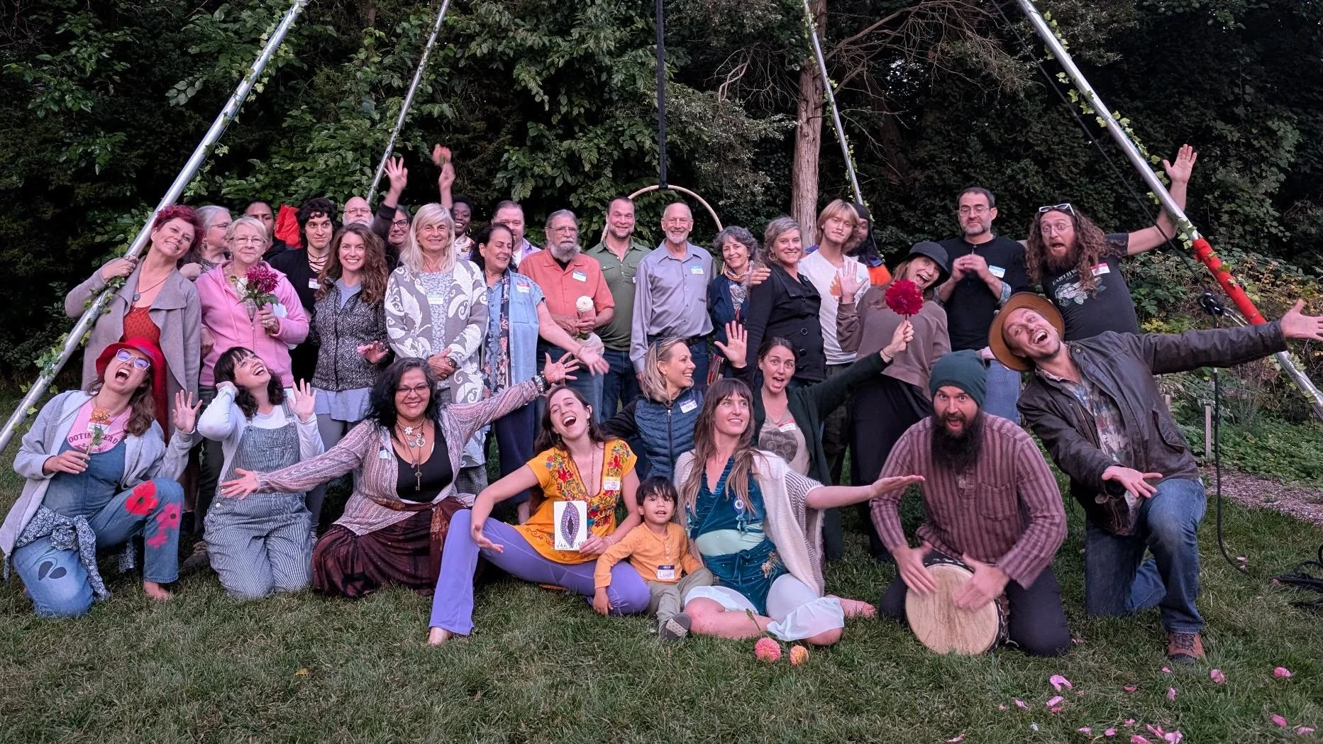 Group of diverse people celebrating outdoors in front of a forested background, smiling, laughing, and waving, with some holding flowers and musical instruments.