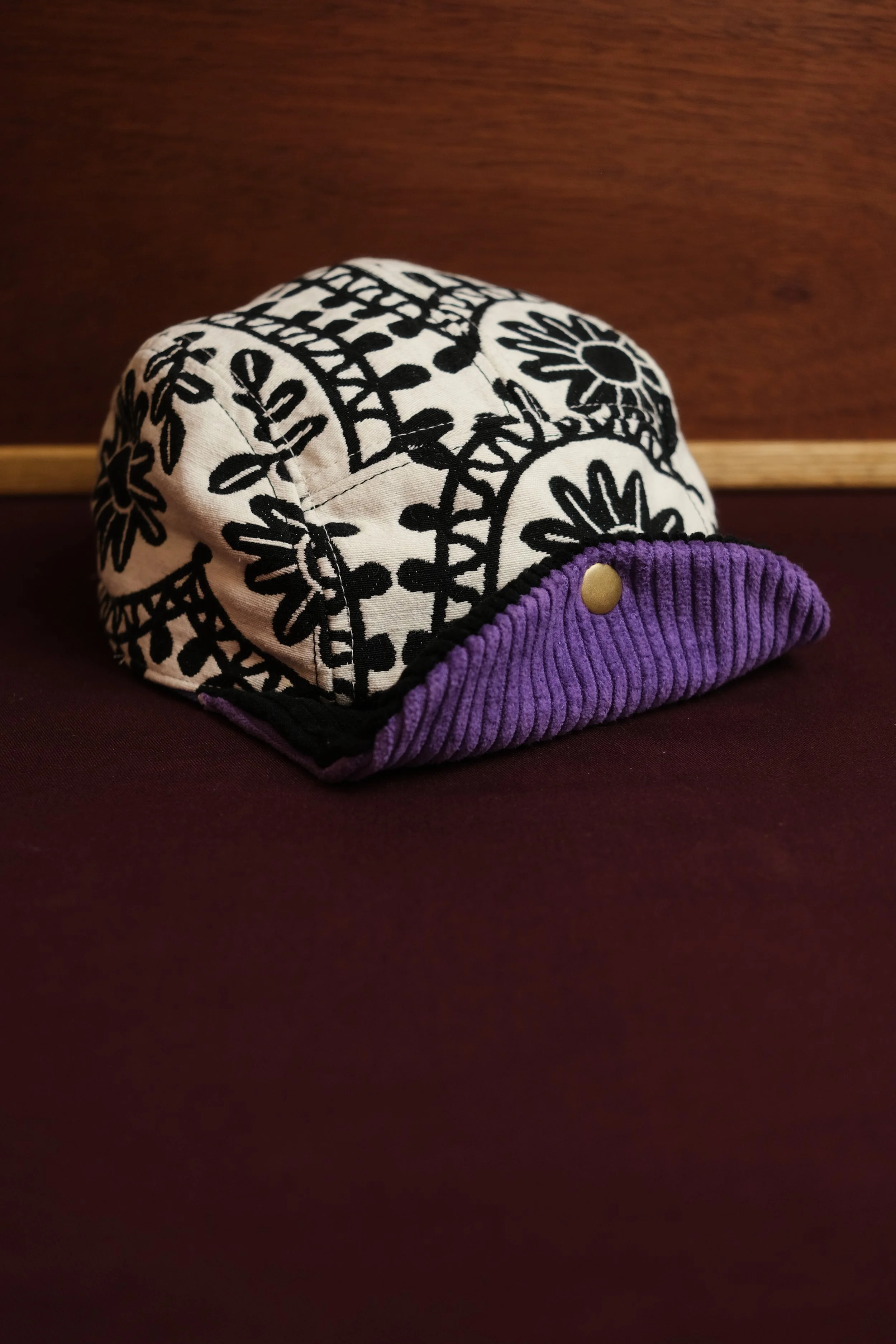 A fabric cap with a black and white floral pattern, purple ribbed brim, and a gold button, placed on a dark surface with a wooden background.