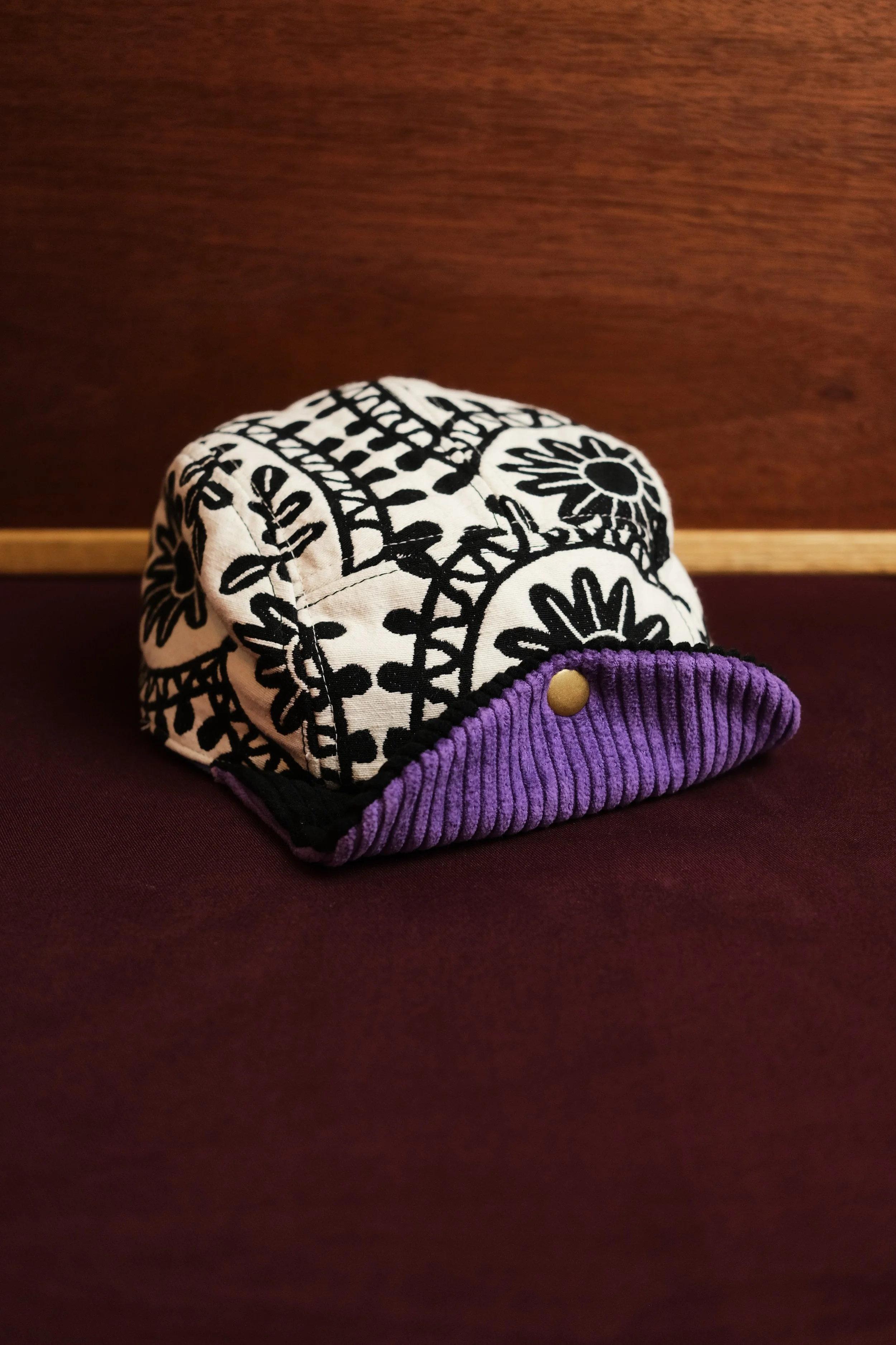 A fabric hat with a black and white floral pattern and a purple ribbed brim, placed on a dark surface against a wooden backdrop.