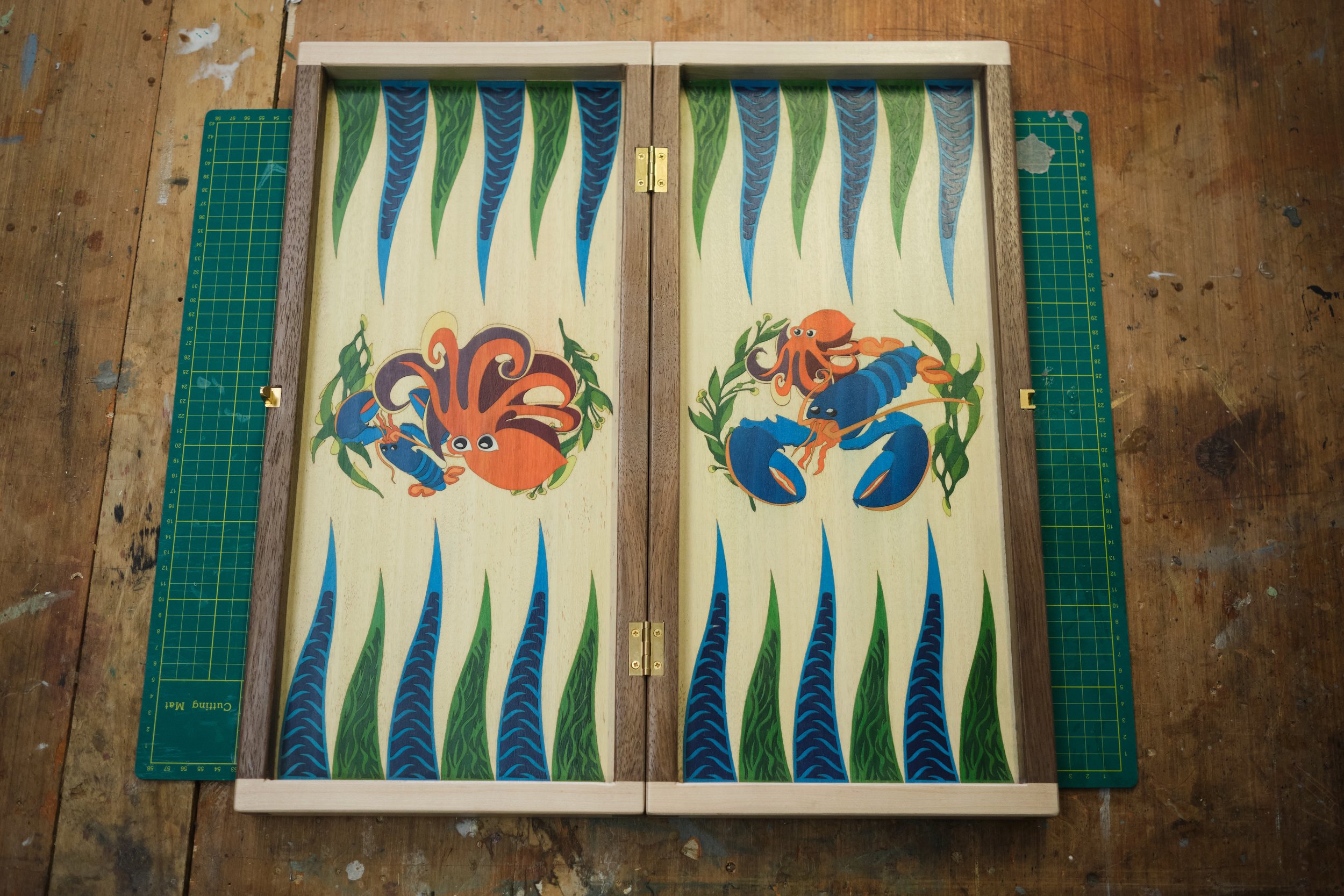 A wooden backgammon board with marine-themed painted illustrations of squid, lobsters, and seaweed, set on a worn wooden surface.