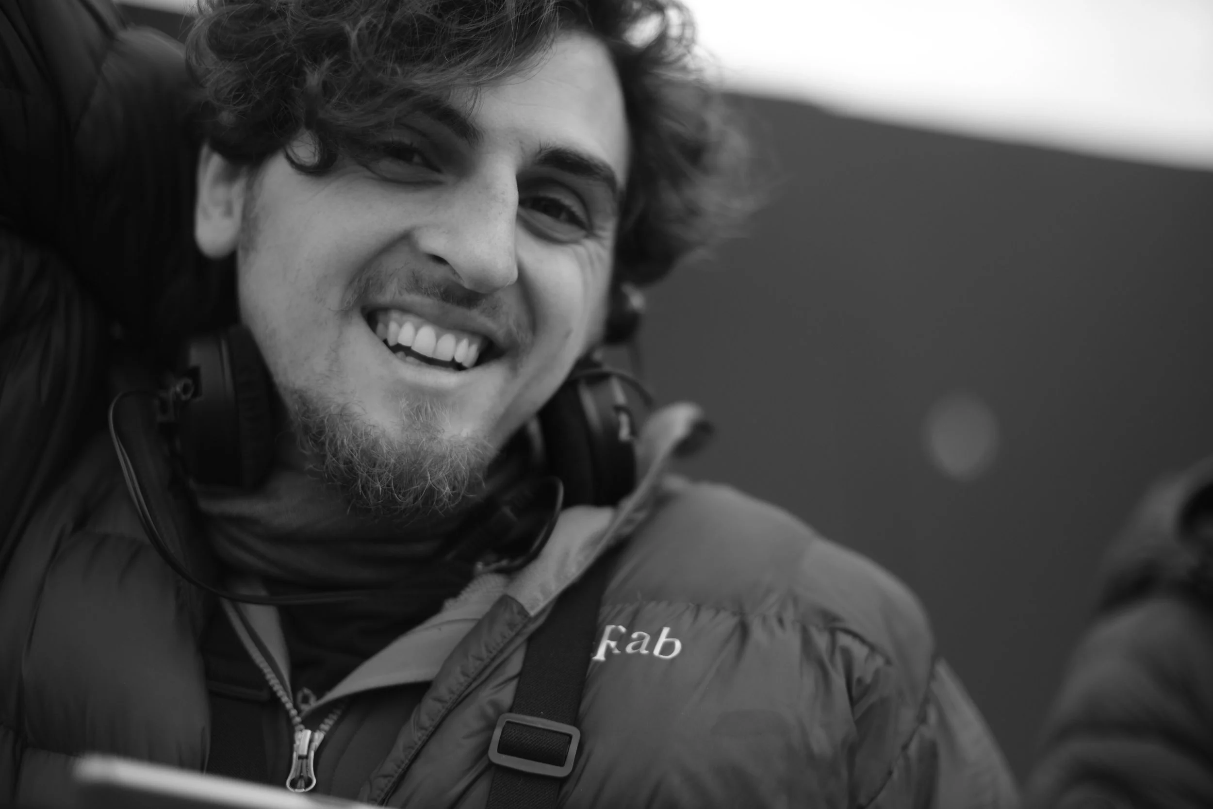 A smiling man with curly hair and a beard looks at the camera. He is wearing a jacket with a visible zipper and a strap across his chest, possibly from a backpack. The background is blurred and appears to be outdoors.