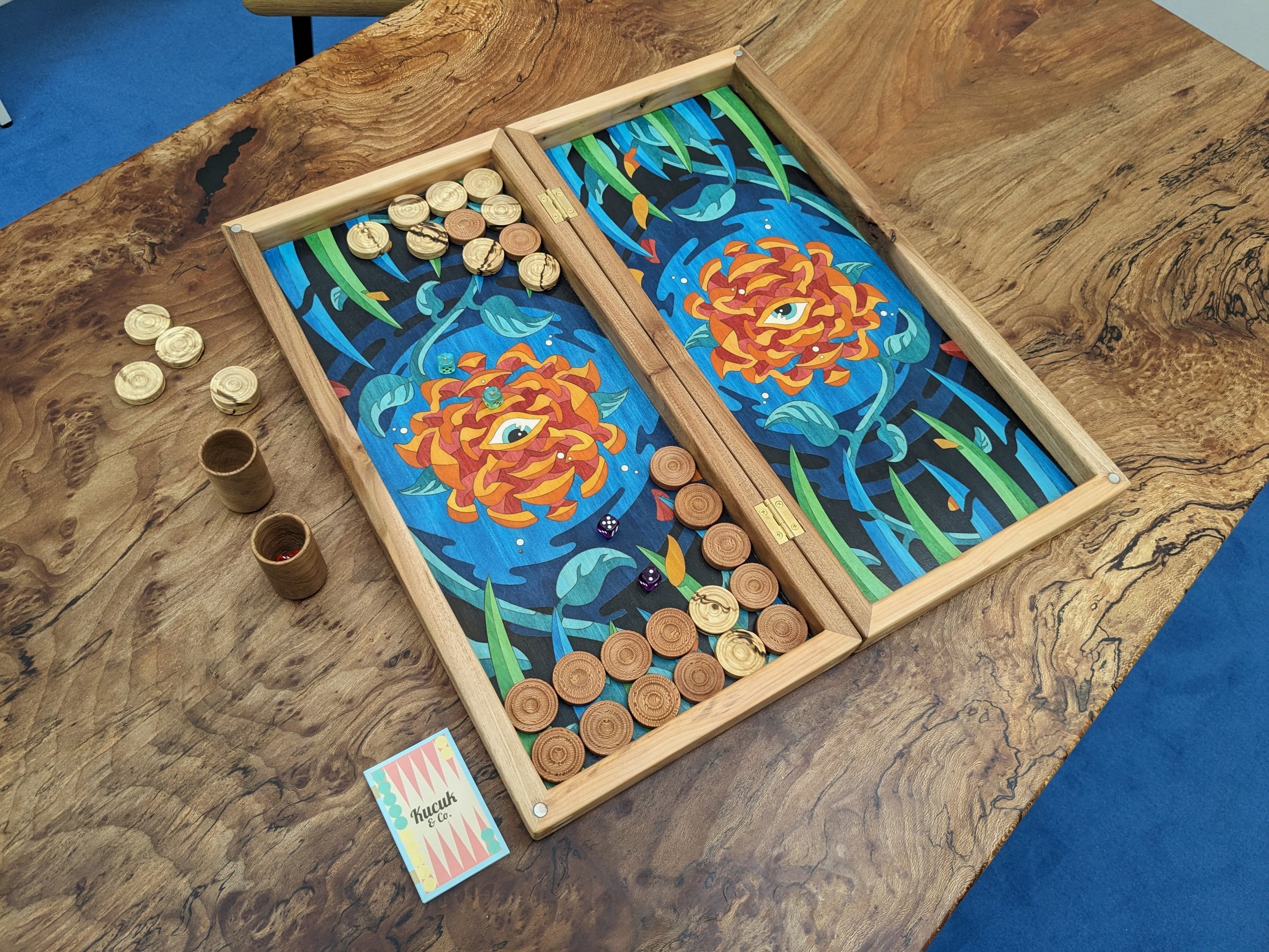 A decorative wooden board game on a wooden table. The game involves moving round wooden pieces, some in a box, with dice rolling on the board. The board features colorful, artistic illustrations of flowers and a background resembling water or sky.