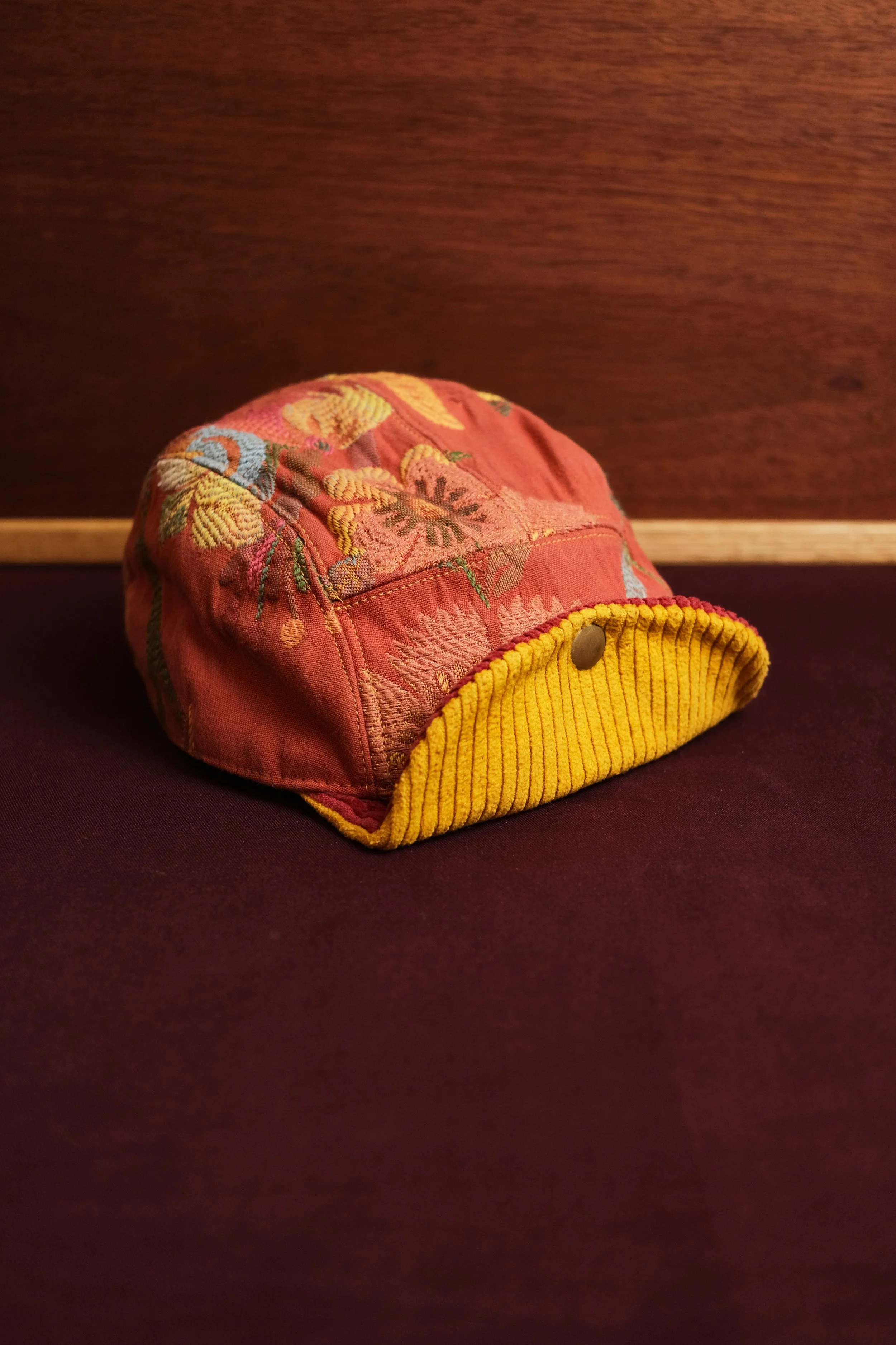 Close-up of a pink quilted hat with embroidered floral patterns, yellow corded edge, and a metal snap button, placed on a dark surface against a wooden background.