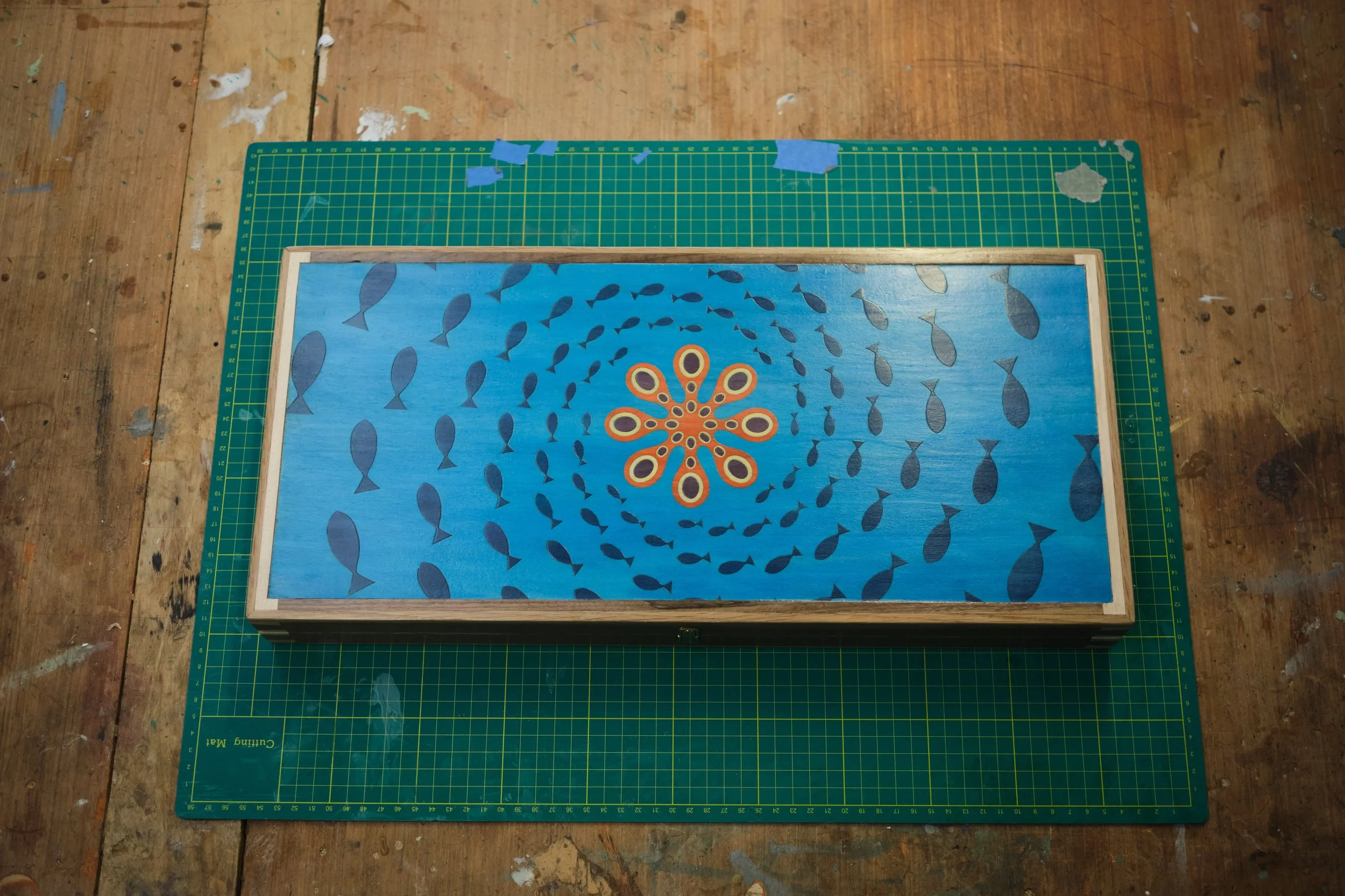 A painted wooden panel with a blue background and a pattern of fish and a flower-like design in the center. The panel is set on a green cutting mat on a wooden work surface with paint splatters and marks.