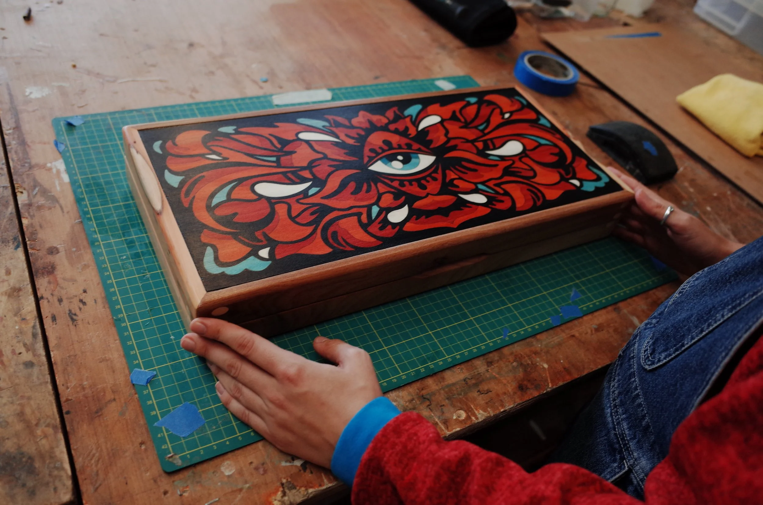 Person holding a framed artwork of a colorful tiger with a blue eye, sitting on a green cutting mat on a wooden table, with art supplies nearby.