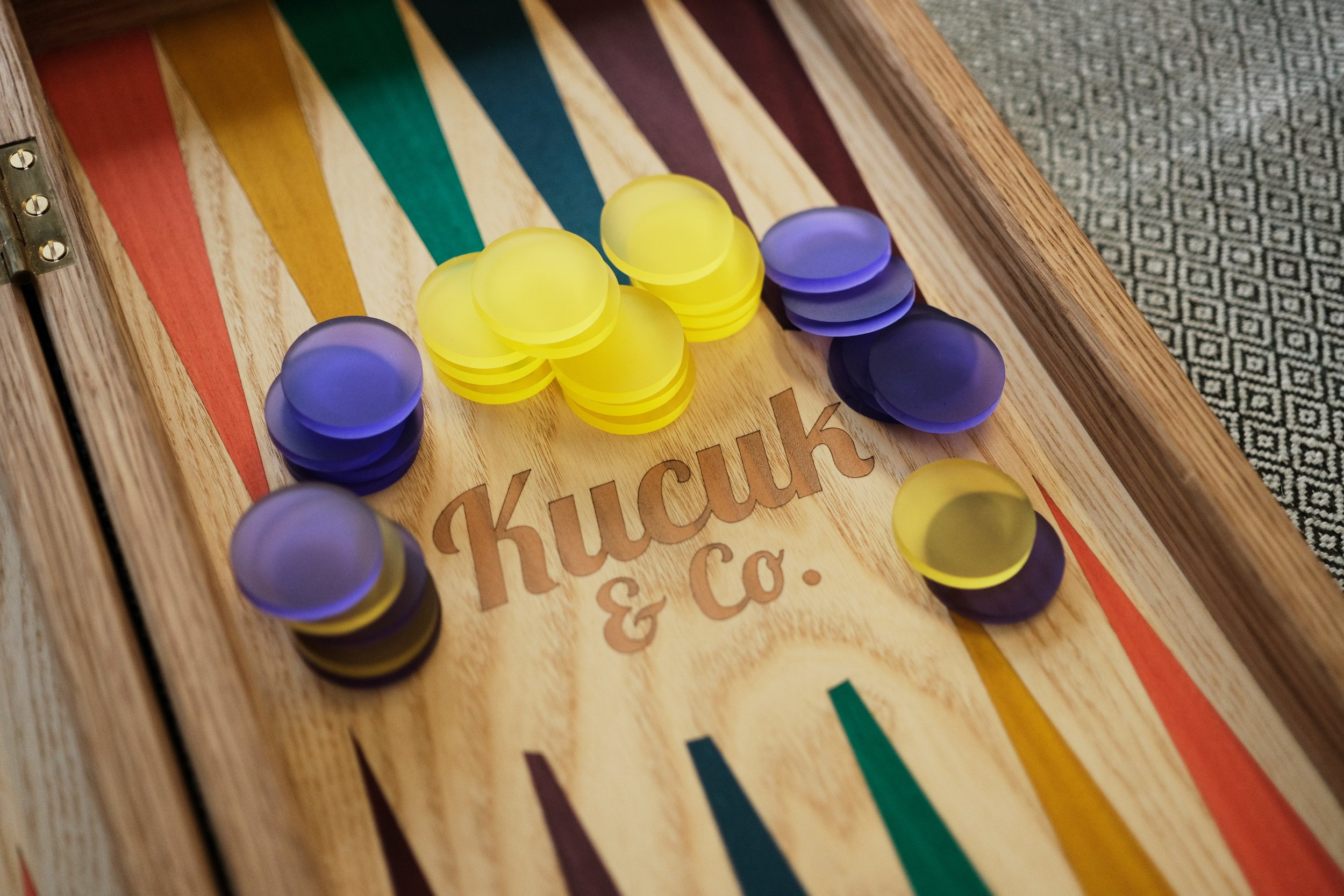 Close-up of a backgammon board with yellow and purple checkers and a wooden surface, with part of a patterned carpet visible on the right side.