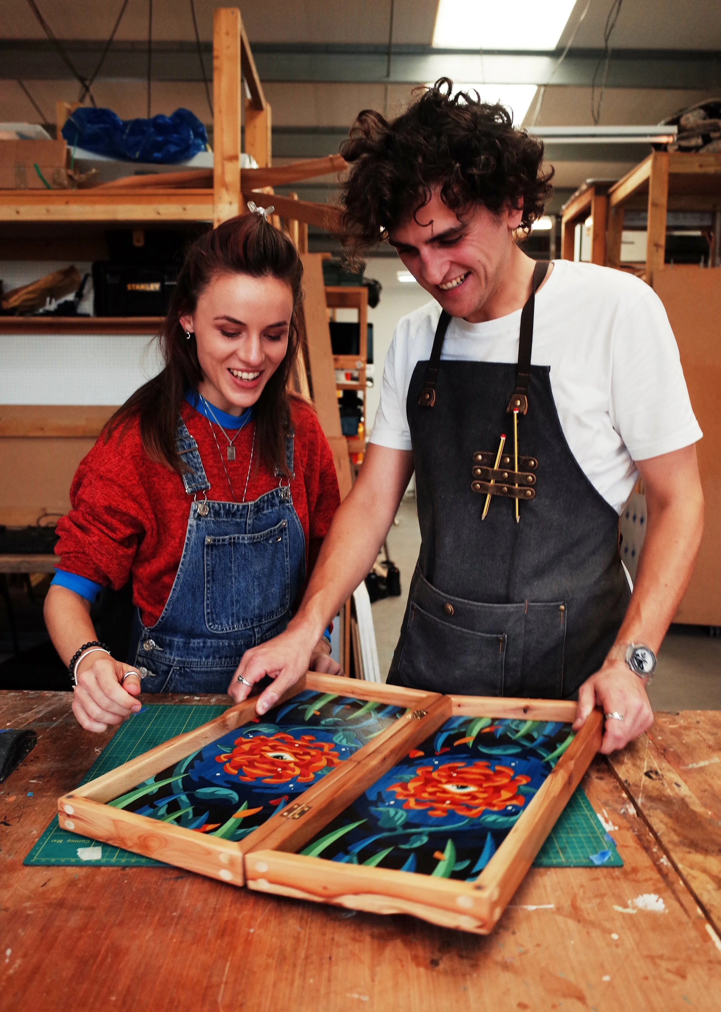 Two young adults, a woman and a man, smiling and working together on a colorful artwork in a woodworking studio. The woman wears a red sweater and denim overalls, while the man wears a white shirt and a black apron. They are standing at a wooden tabl
