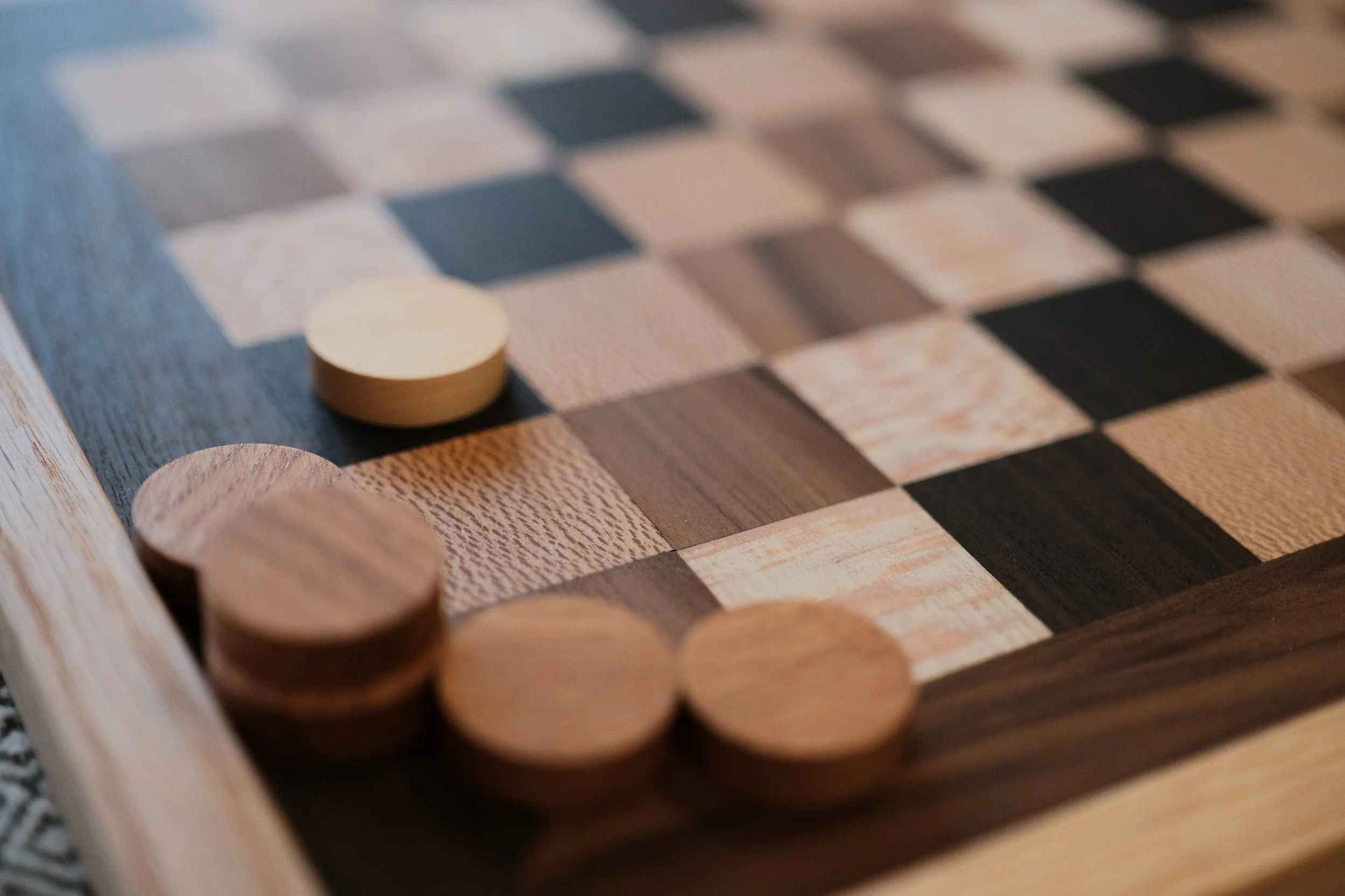 Close-up of a wooden chessboard with black and beige squares, with several round wooden chess pieces, some in focus, placed on the board.