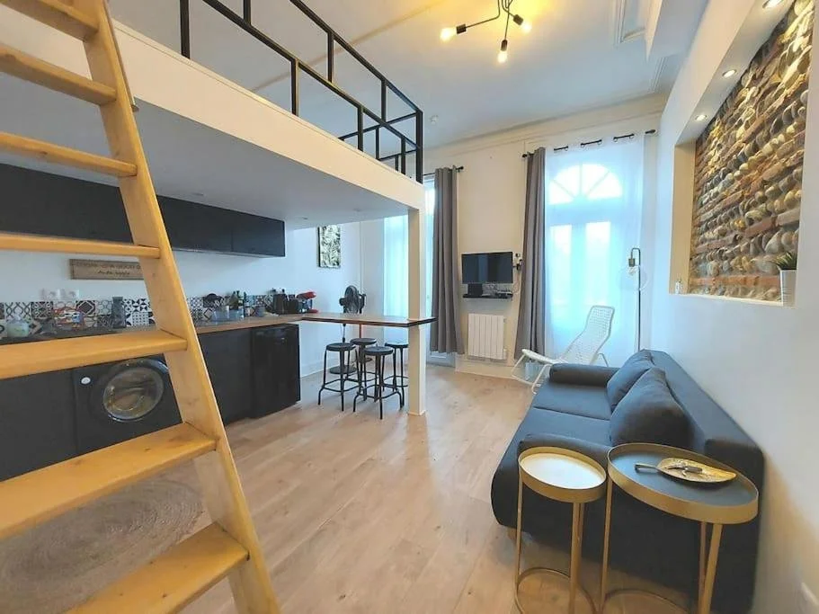 Salon spacieux avec canapé gris, deux petites tables rondes, cheminée en pierre, télévision, canapé blanc près d'une grande fenêtre, marche en bois menant à une mezzanine.