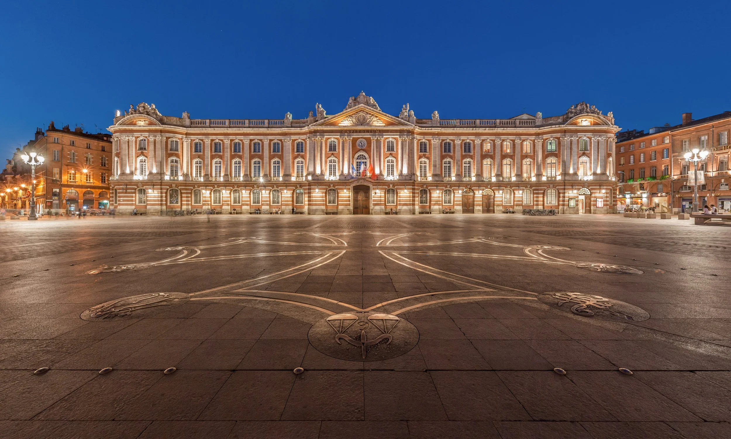 Bâtiment historique éclairé la nuit dans une place pavée avec des motifs en métal, entouré d'immeubles résidentiels, sous un ciel bleu foncé.