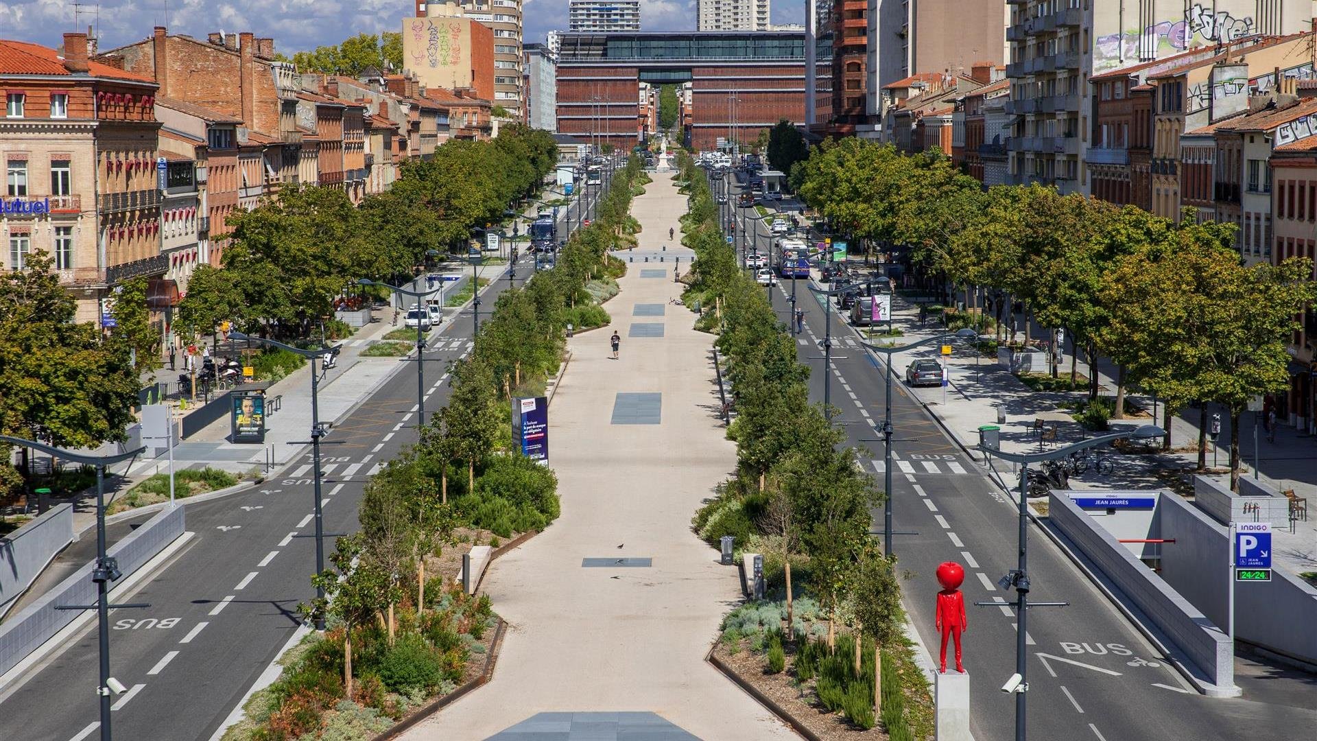 Vue aérienne d'une rue urbaine avec des bâtiments résidentiels et commerciaux, une voie piétonne centrale bordée d'arbres, et des voitures stationnées et en mouvement.