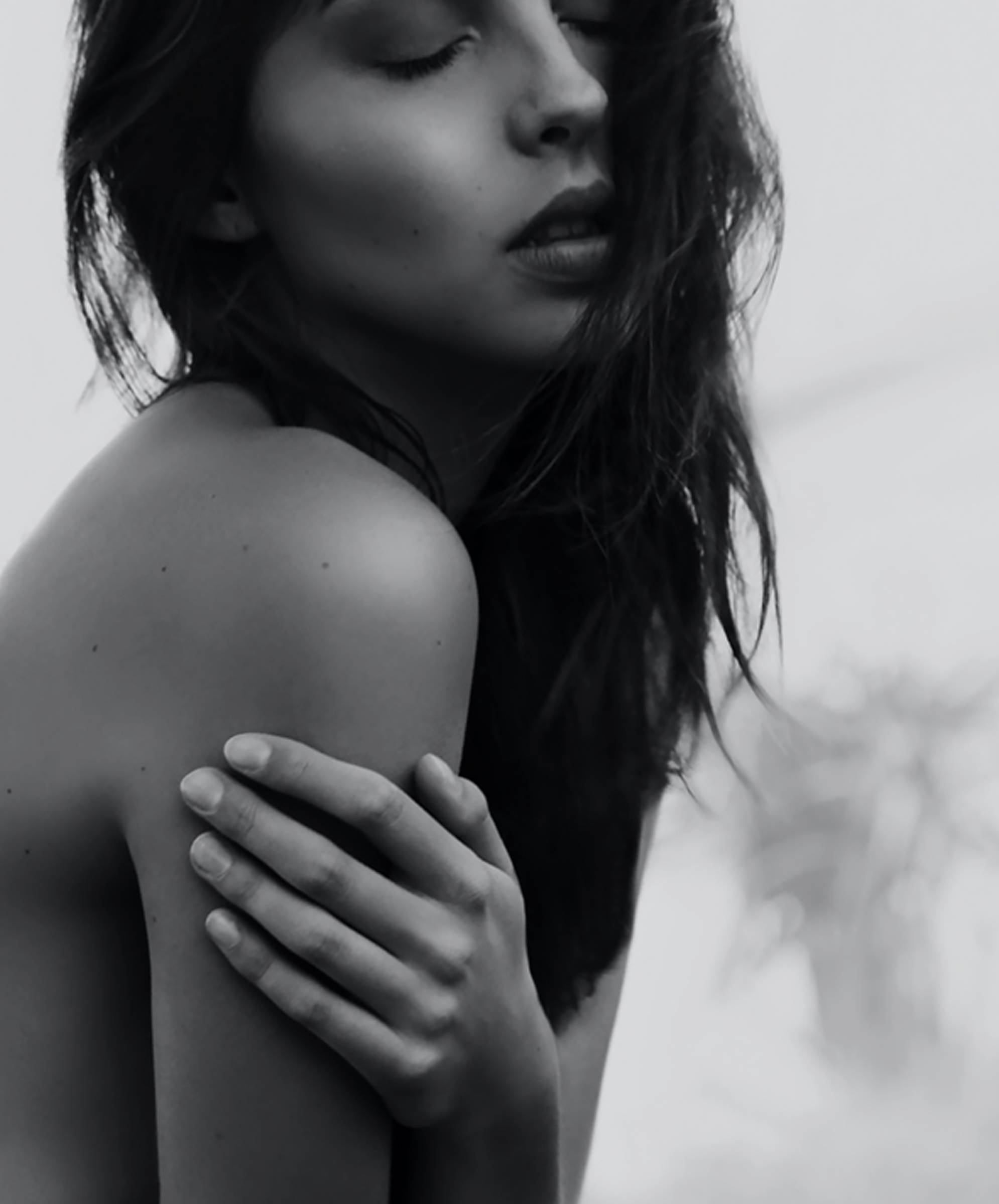 A close-up black and white photograph of a woman with long hair, her eyes closed, lips slightly parted, and one hand resting on her shoulder.