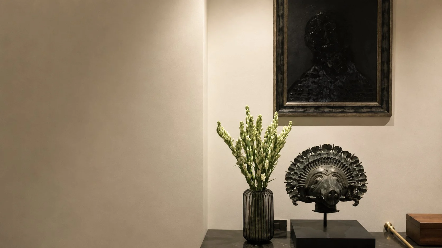 A vase with tall white flowers, a carved metal mask resembling a hog, and a framed dark portrait on a wall.