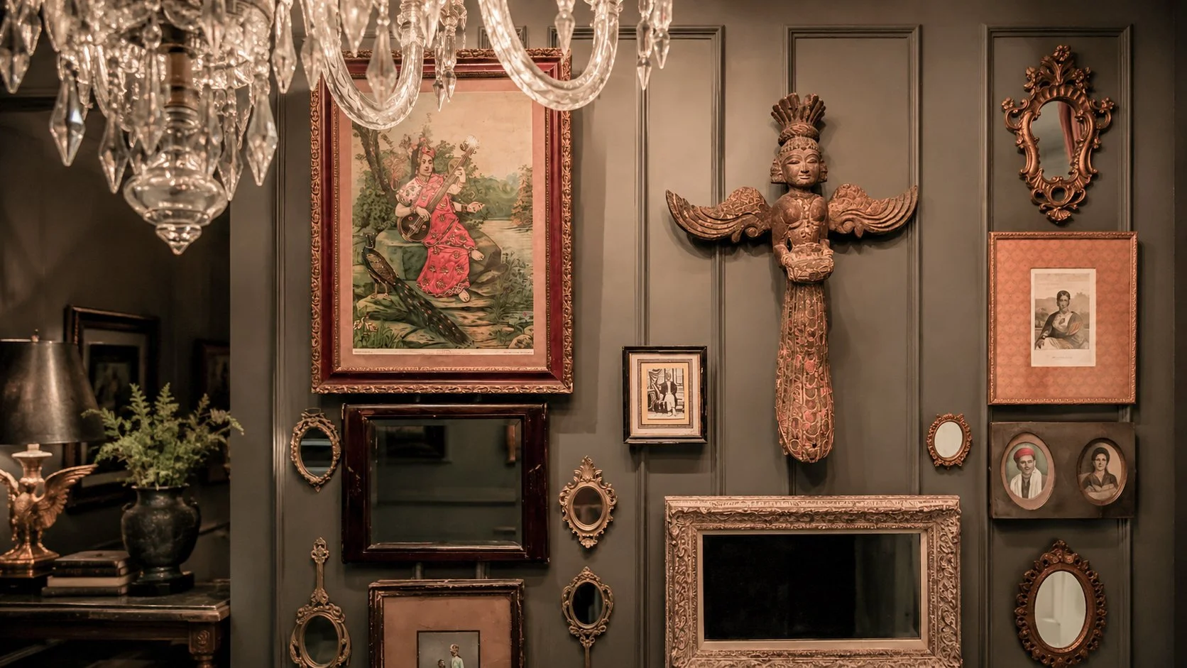 Decorative gallery wall with ornate picture frames, a carved wooden tribal mask, paintings, and photographs in a dimly lit room. A chandelier with crystal pendants hangs overhead, and a table with a dark vase and greenery is visible on the left.
