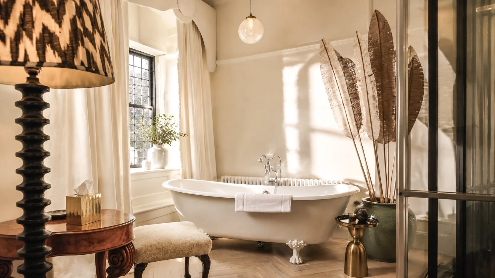 A bathroom with a vintage white clawfoot bathtub near a window with curtains, decorated with dried leaves in a large pot, and a glass shower enclosure to the right. A lamp, tissue box, and a stool are also visible.