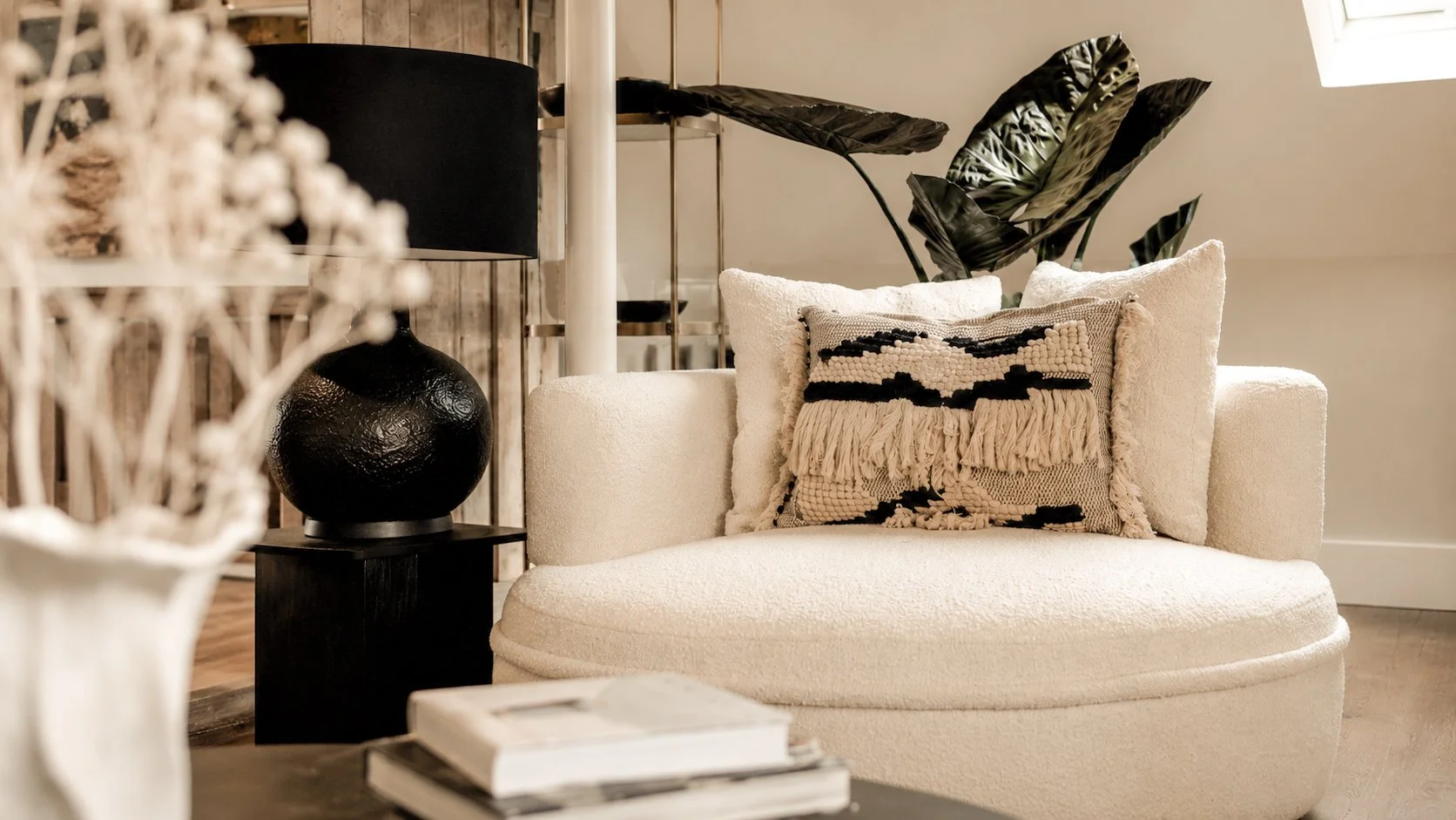 Interior of a living room with a white curved sofa, decorative pillows including a textured one, a black table lamp with a black shade, a dark side table, a modern plant with large dark leaves, and a window allowing natural light to enter.