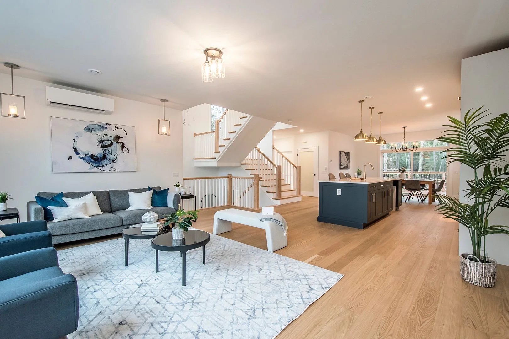 Open living space with gray sofa, blue armchairs, coffee tables, abstract wall art, staircase, modern kitchen with island, dining table, large window, and indoor plants.