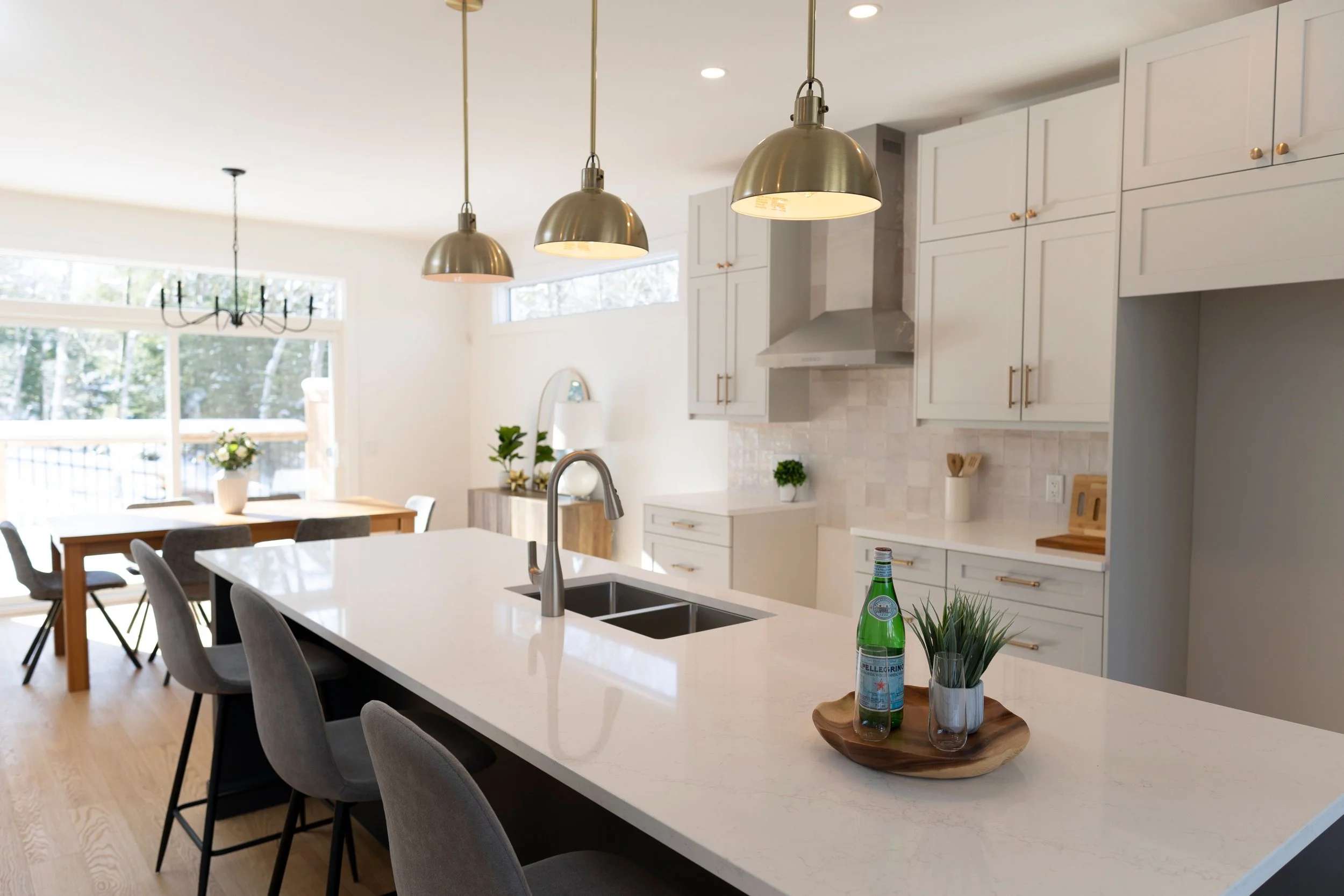 Modern open-concept kitchen and dining area with a white island, gray chairs, white cabinets, and gold accents. Three pendant lights hang over the island, and a large window with natural light and trees outside is visible.