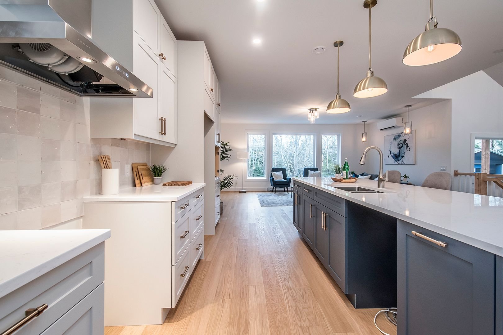 Open plan kitchen with white upper cabinets, gray lower cabinets, a large island with a sink, pendant lights, and a living room with large windows and seating in the background.