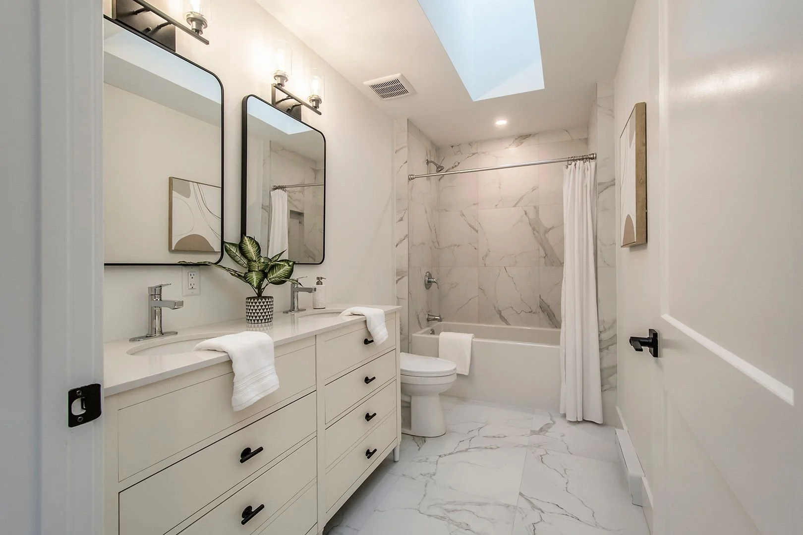 A modern bathroom with a white double vanity, black handles, two oval mirrors, a potted plant, and a white bathtub with marble walls and a shower curtain.