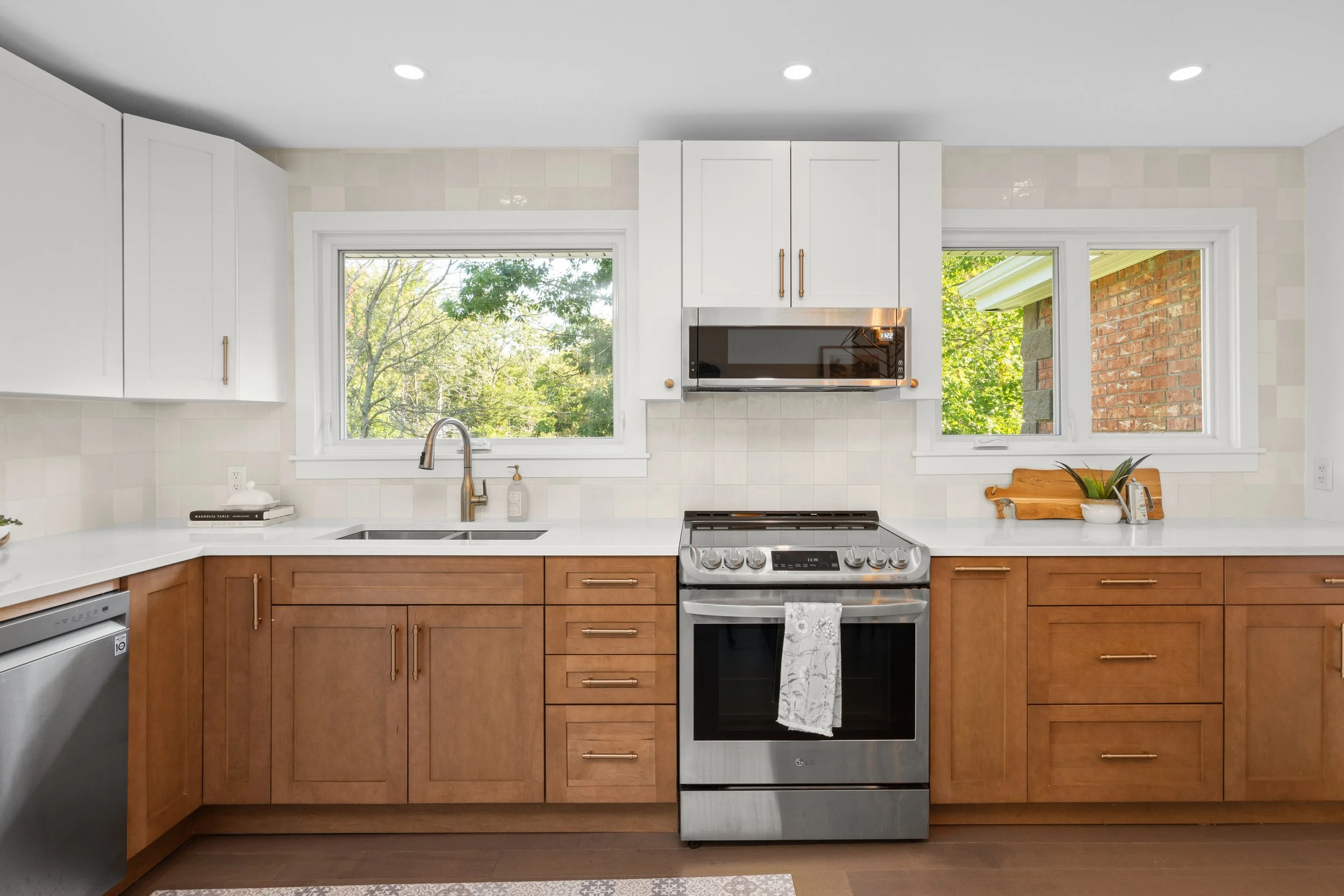 A kitchen with white upper cabinets, wooden lower cabinets, a window showing trees outside, a microwave above a stove, and a small countertop with a pineapple and a cutting board.