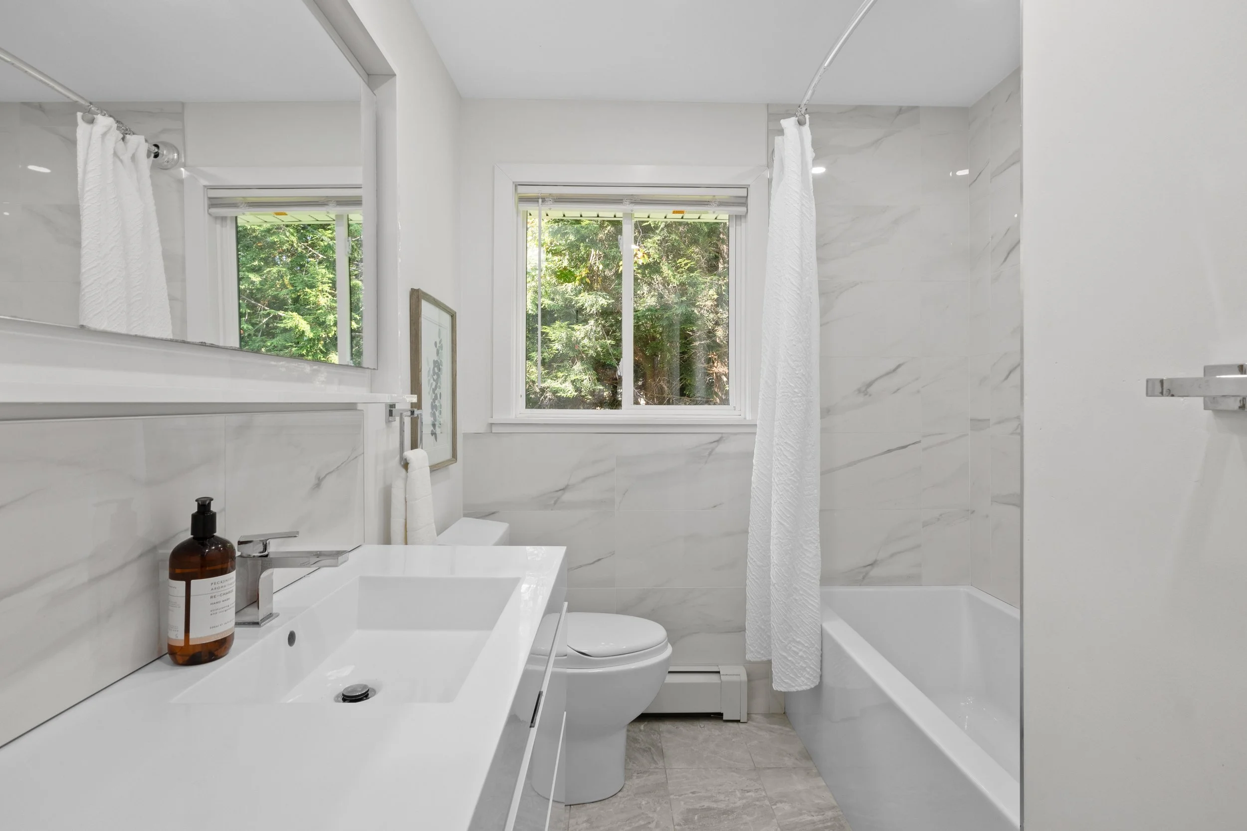 Clean modern bathroom with white marble walls and floor, window showing green trees outside, white fixtures, a mirror, and a bottle of soap on the sink.