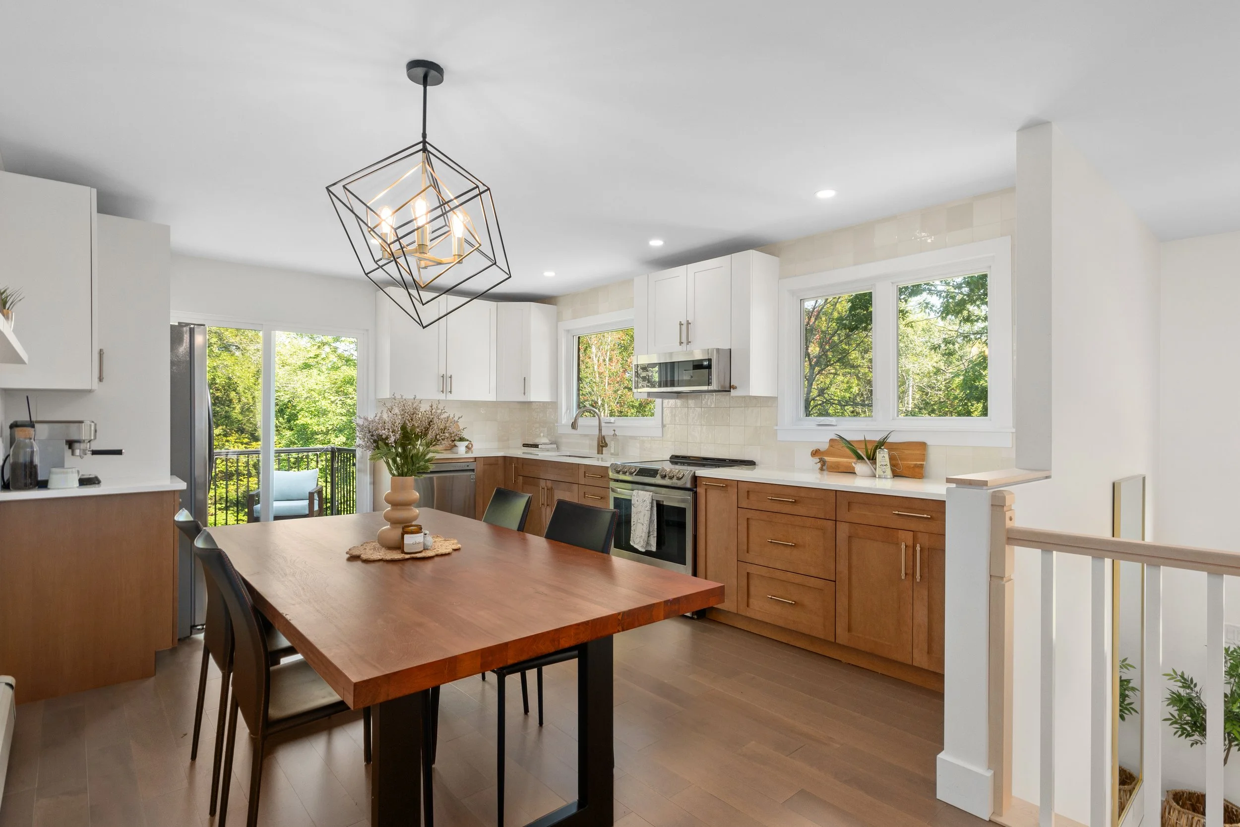 A modern kitchen with white cabinets, wooden lower cabinets, stainless steel appliances, and large windows showing green trees outside. There is a wooden dining table with black chairs, a vase with flowers, and a contemporary geometric chandelier han