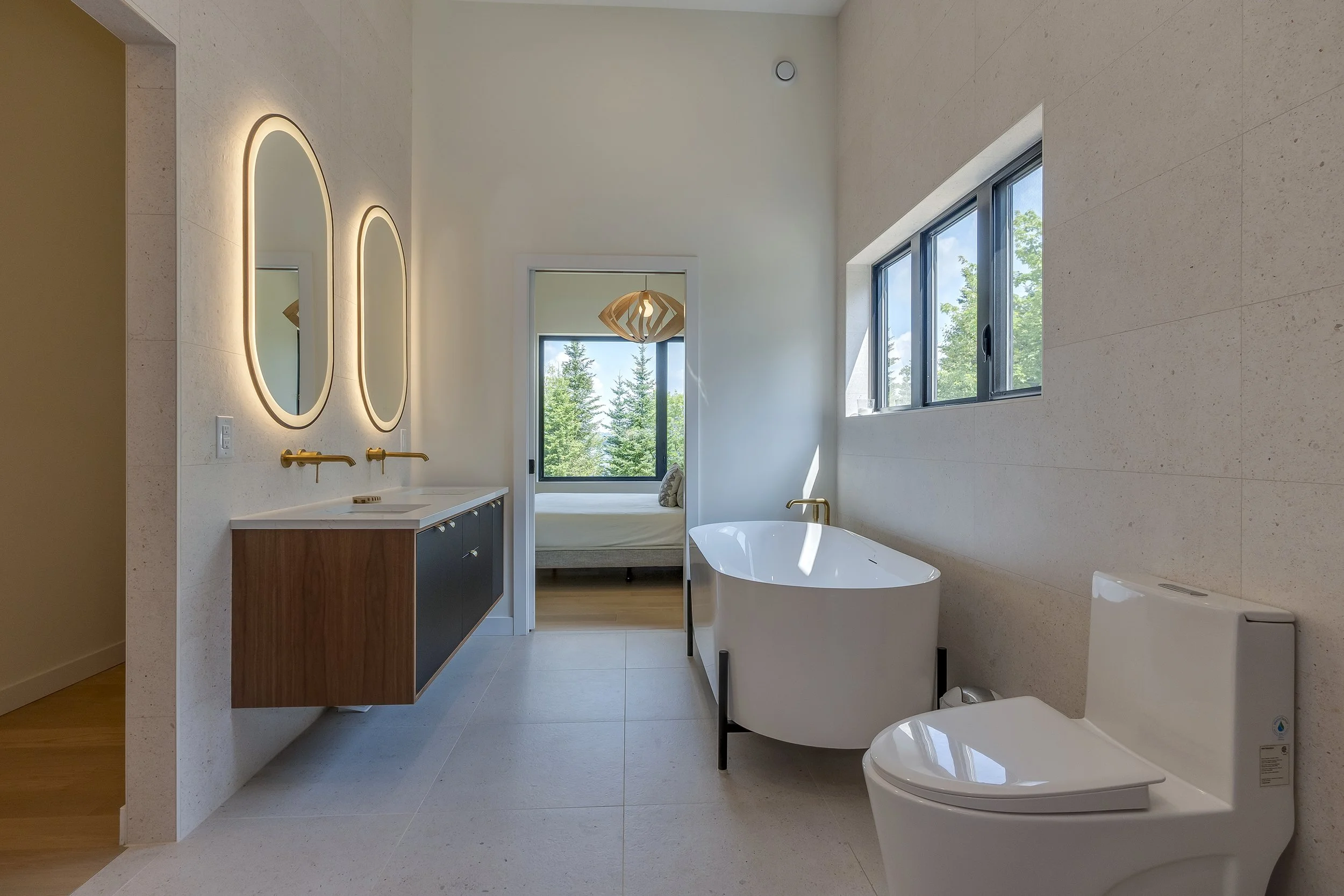 Modern bathroom with a white bathtub, a toilet, a dark vanity with a white countertop, and two oval mirrors with backlit lighting. Large windows show a view of green trees and blue sky.