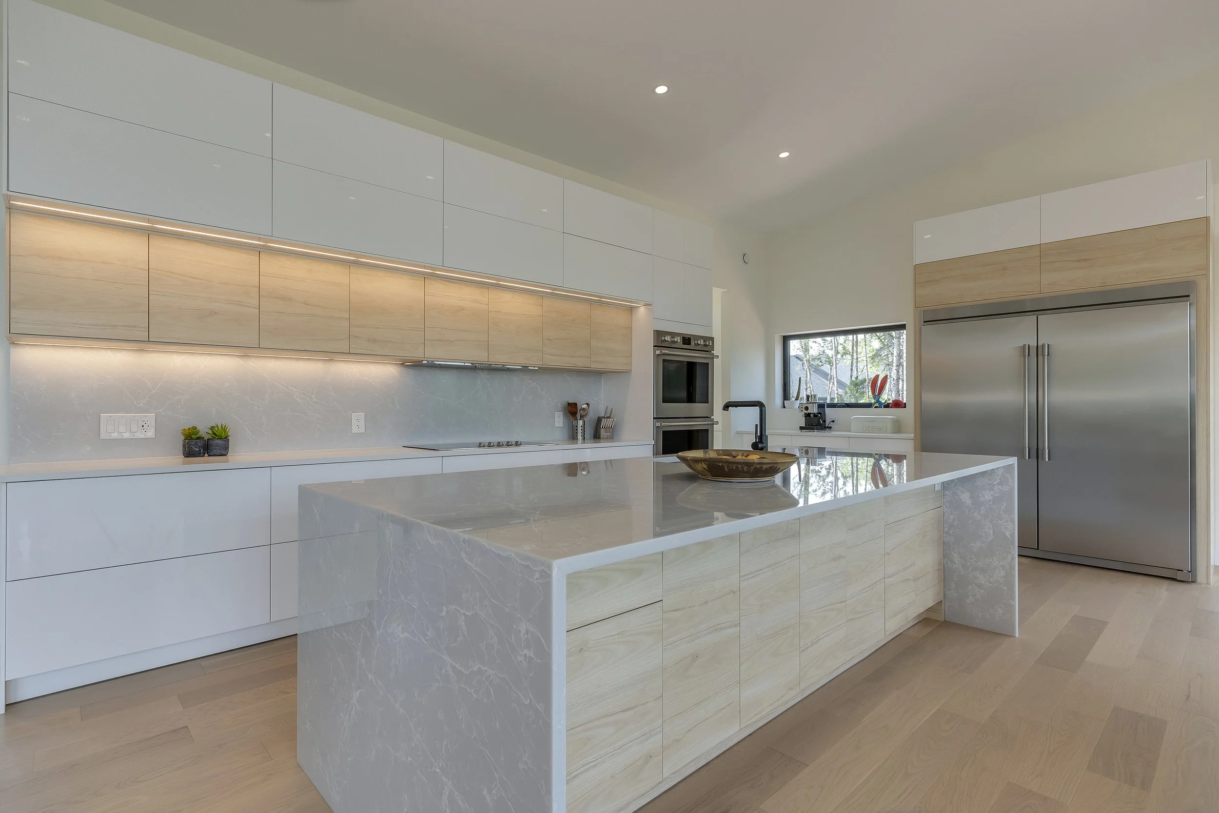 Modern kitchen with white and light wood cabinets, marble island, stainless steel refrigerator, oven, black faucet, and small potted plants, with a window showing trees outside.