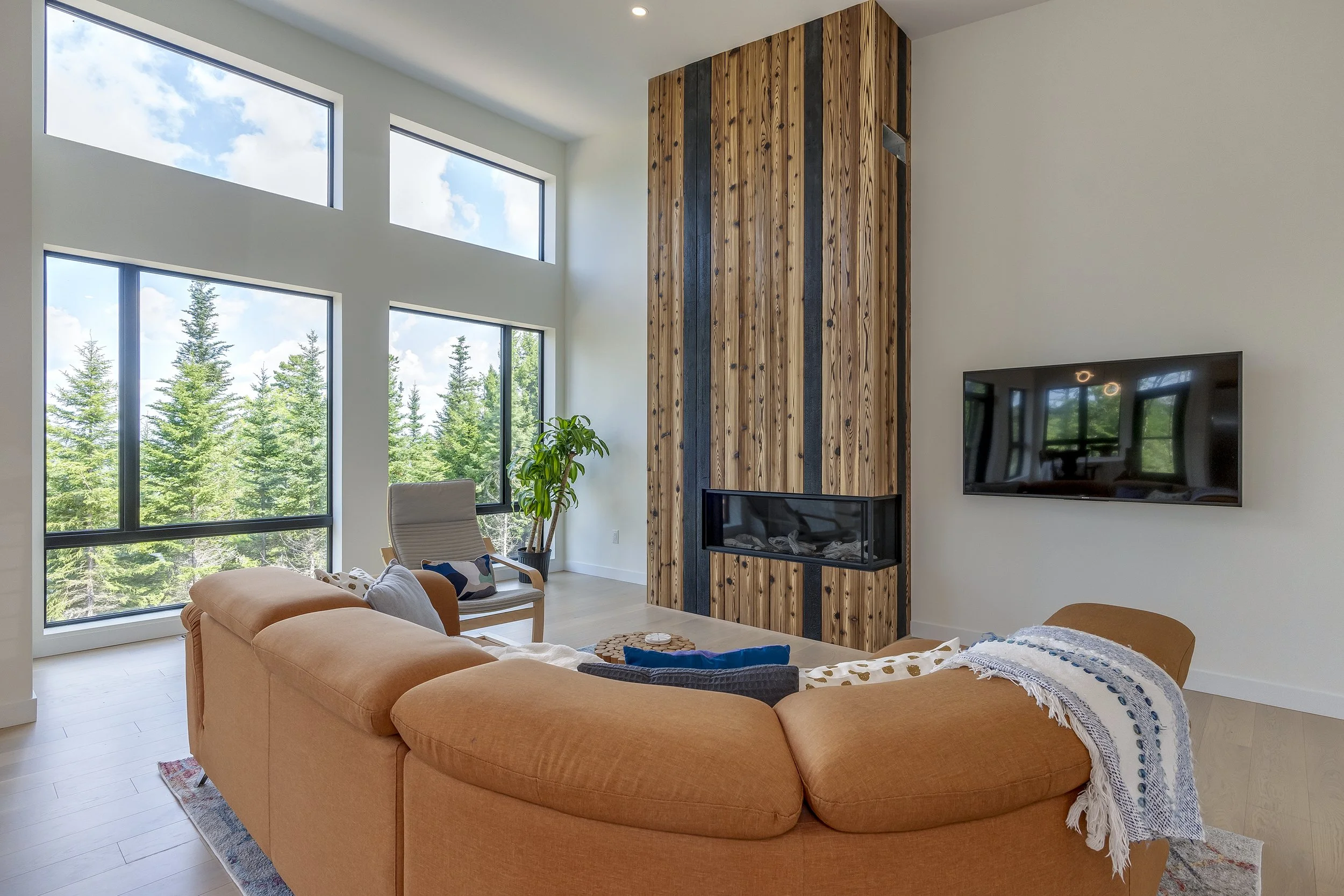 Modern living room with large windows, an orange sectional sofa, a beige armchair, a potted plant, and a wood panel fireplace with black accents.