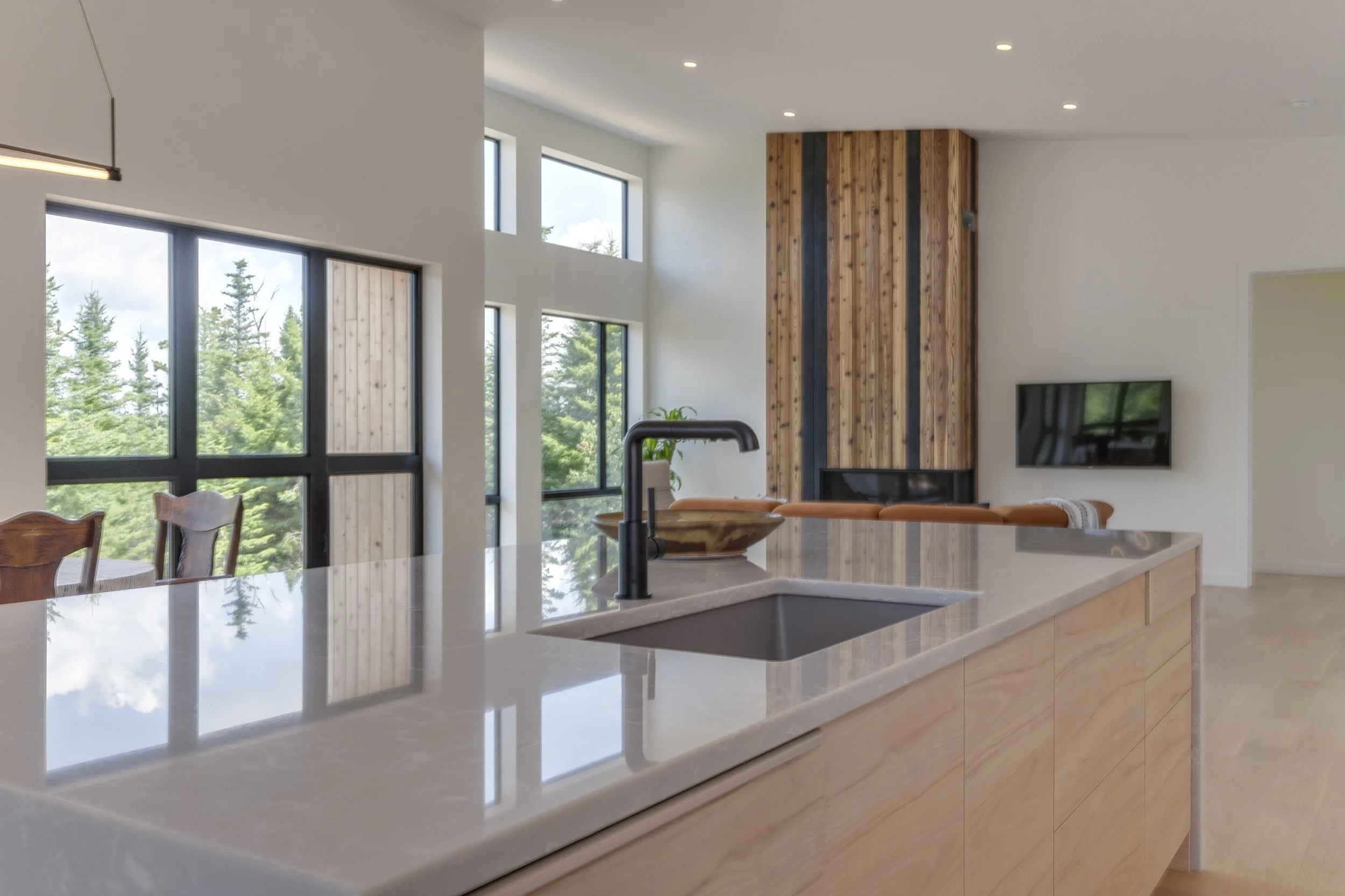 Modern kitchen with a white countertop, a black sink faucet, and a large window showing trees outside. A TV is mounted on a wooden-paneled wall near the ceiling, and there are wooden chairs around a dining table.