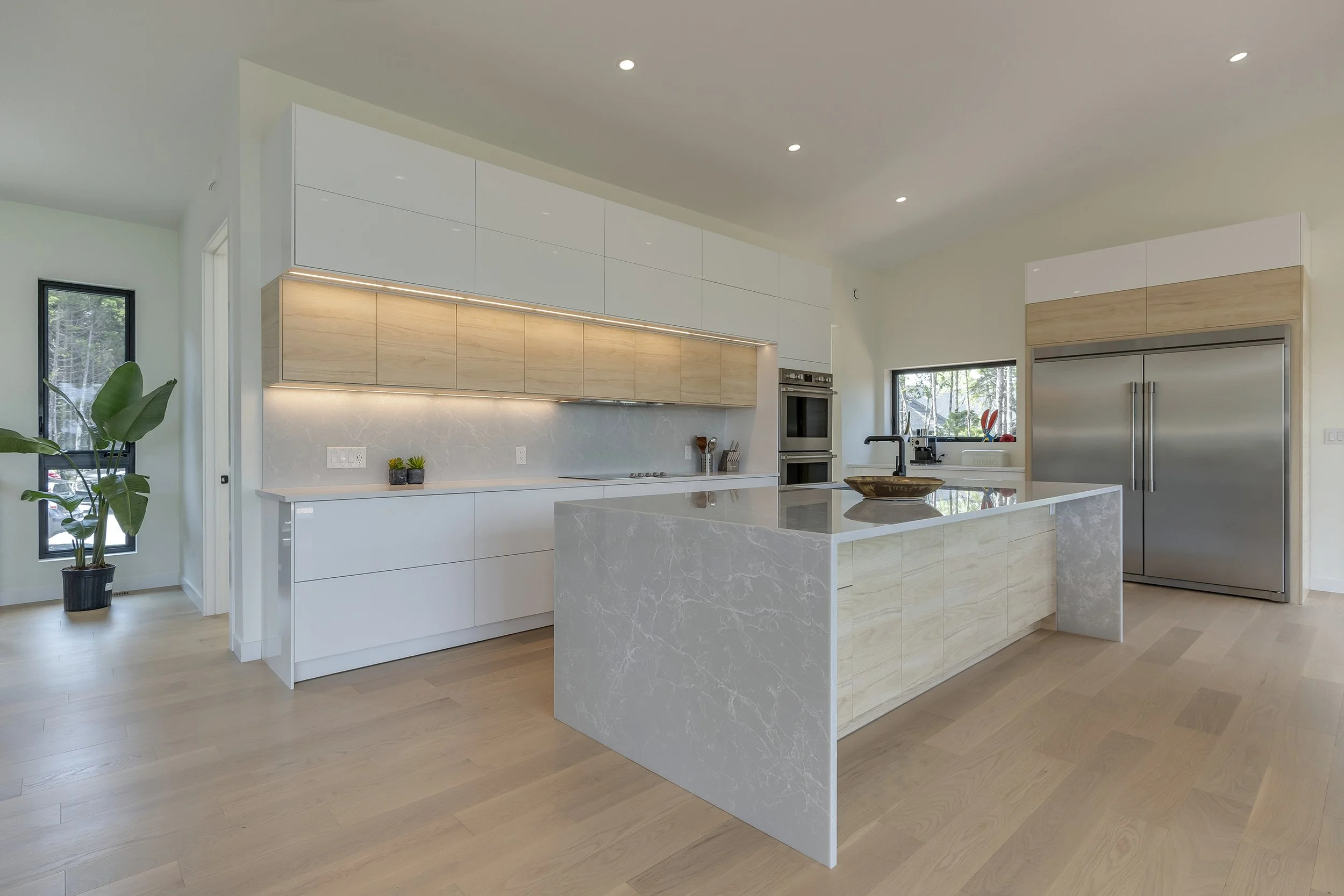 Modern kitchen with white cabinets, a marble island, stainless steel refrigerator, and light wood accents. Large windows with a view of greenery and a potted plant in the corner.