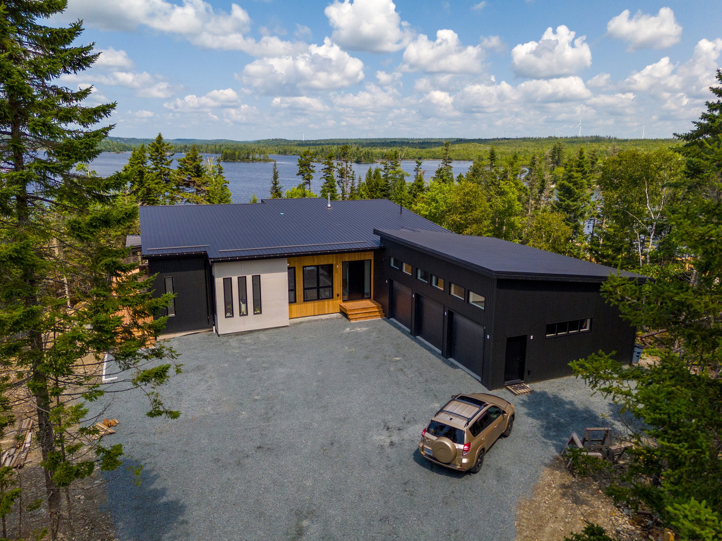 A view of a modern building with black and white exterior walls and wooden accents, situated in a natural setting with trees and overlooking a body of water, under a blue sky with scattered clouds.
