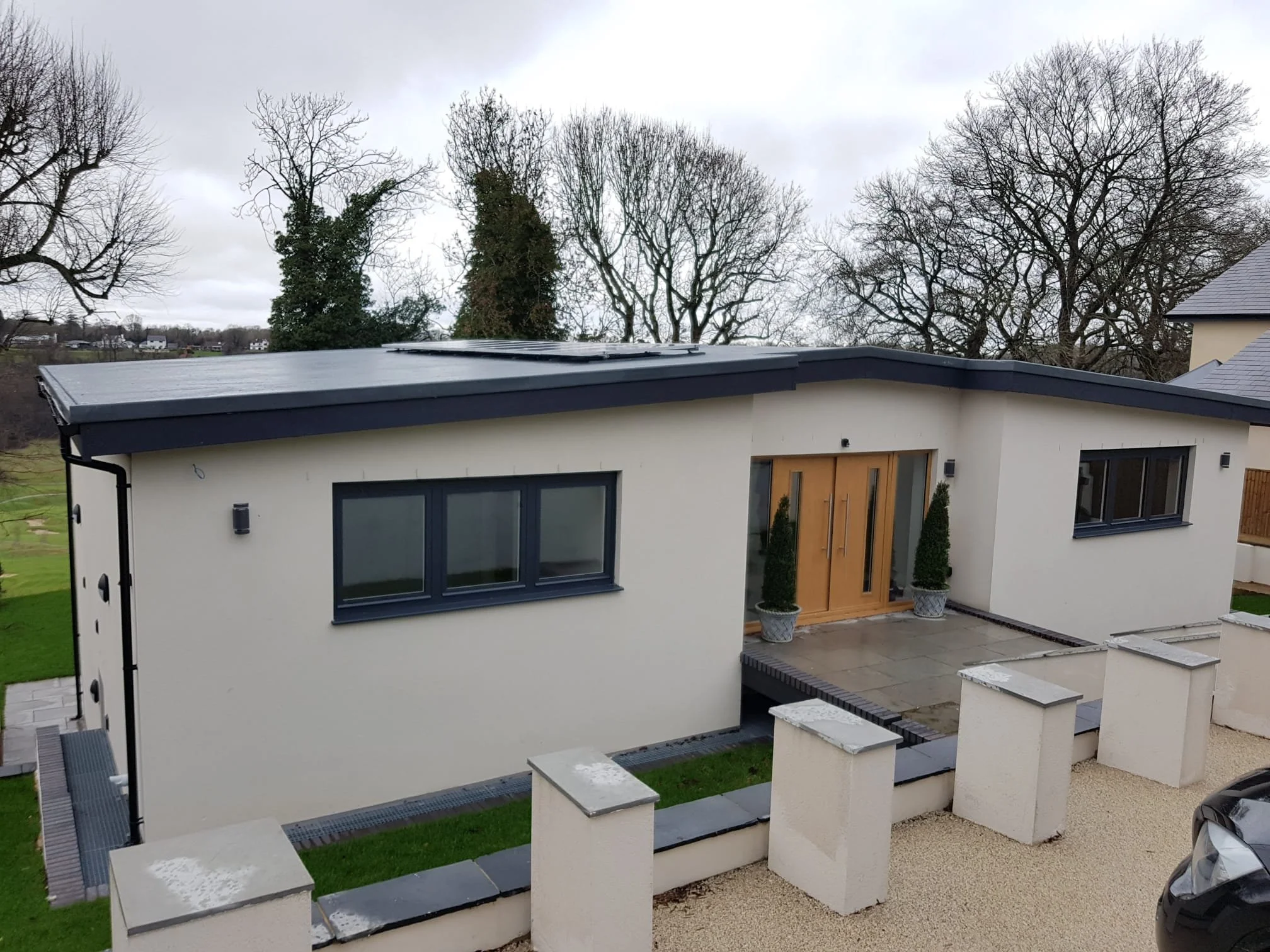 Modern house with flat roof and large windows, surrounded by trees and a landscaped yard with stairs and planters.