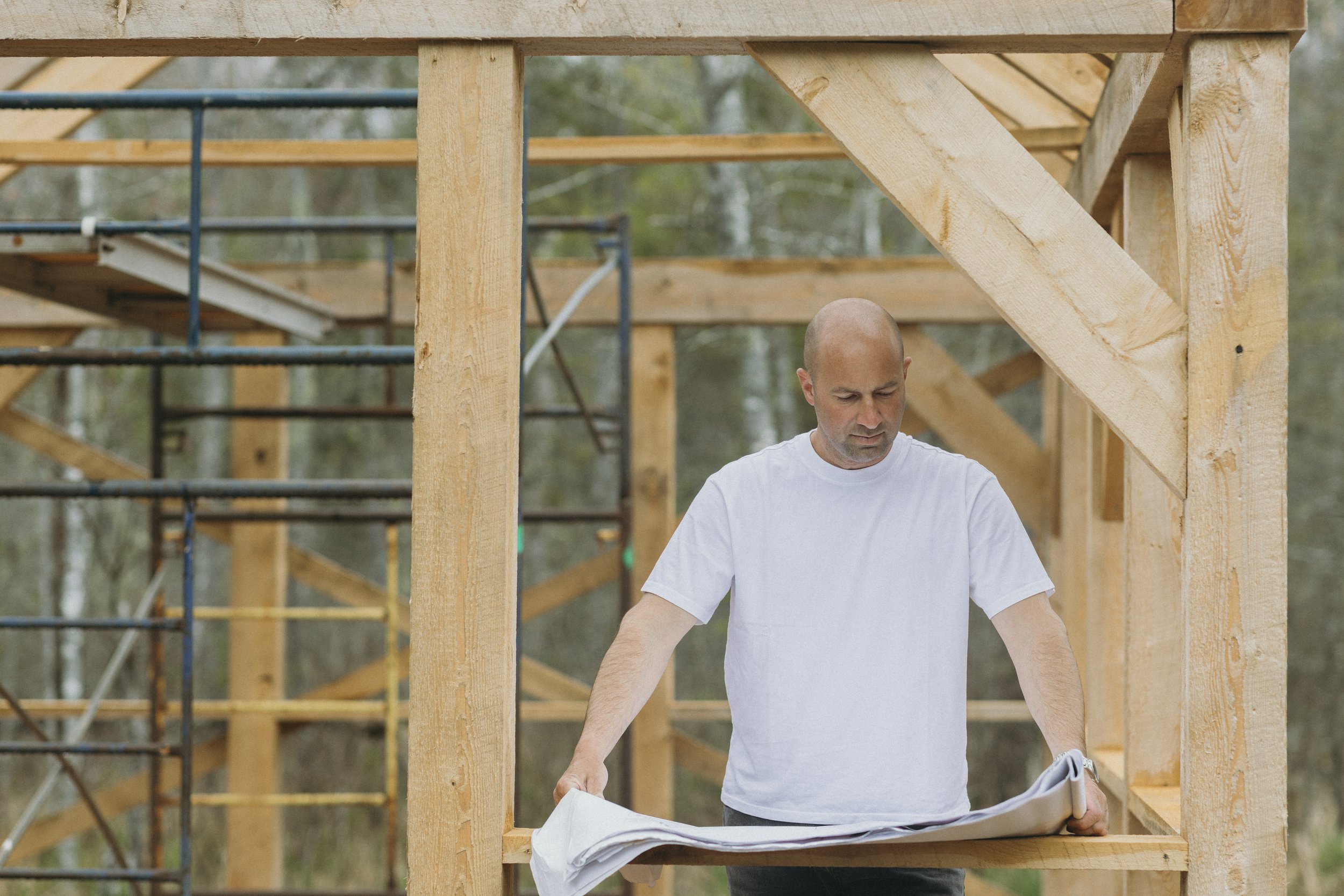 Mark Bemister in a white t-shirt working on a wooden structure in an outdoor setting with trees in the background.