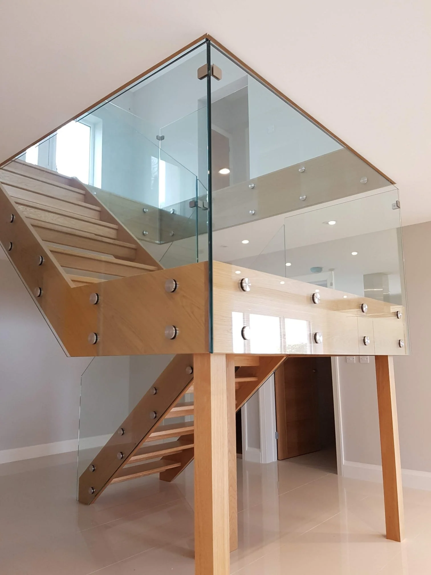 Interior view of a modern staircase with wooden steps, glass railing, and a landing that leads to a room with white walls and beige tiled flooring.