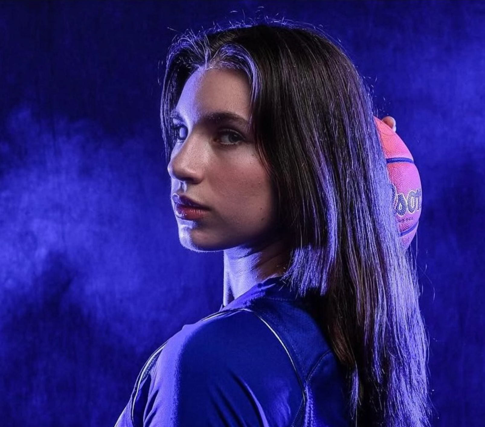 A woman with wet hair and a serious expression, wearing a dark jacket, stands in front of a dark blue background with purple lighting.