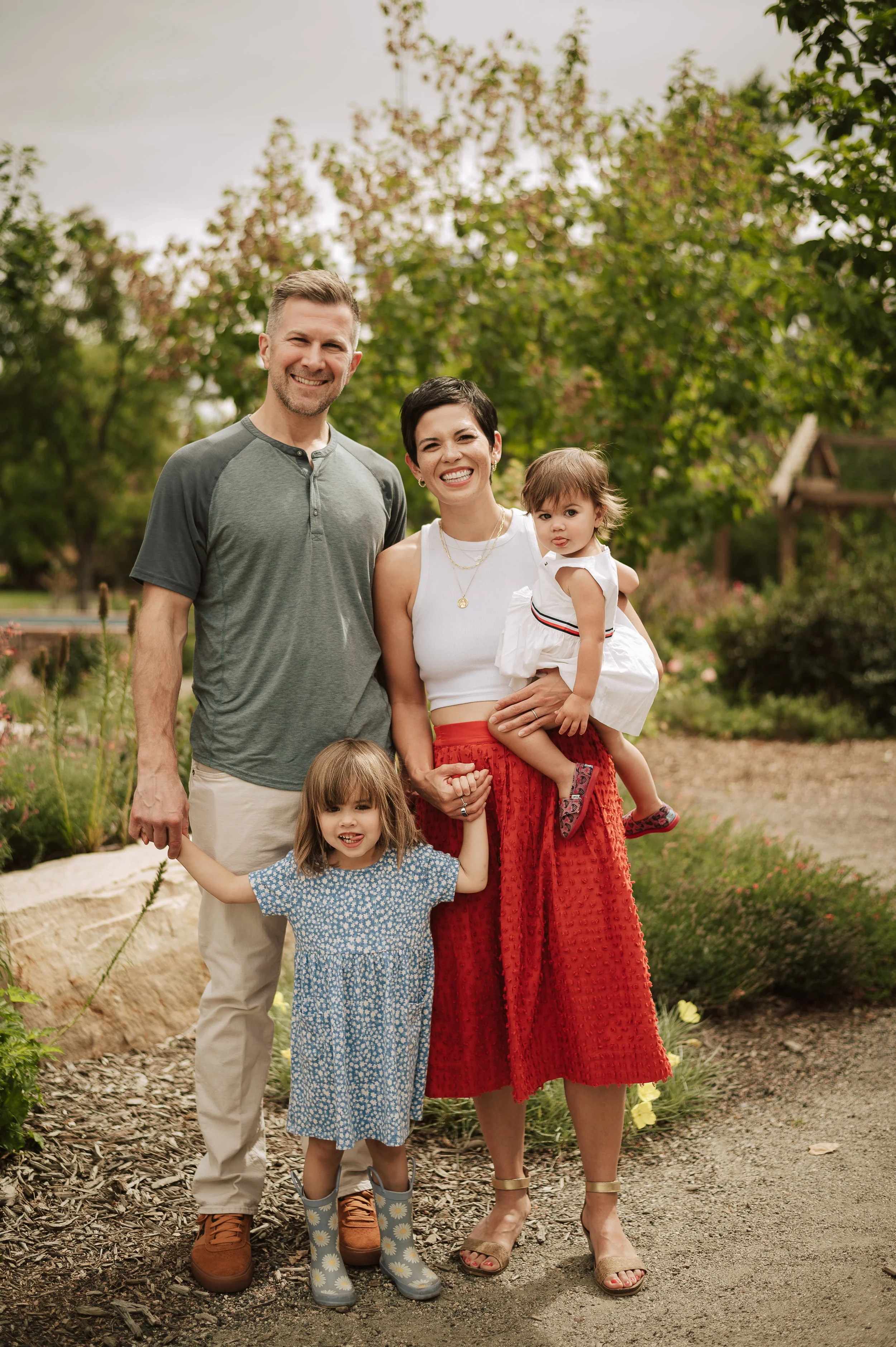 A smiling family of four outdoors, including a man, woman, and two children, standing on a garden trail with trees and flowers in the background.
