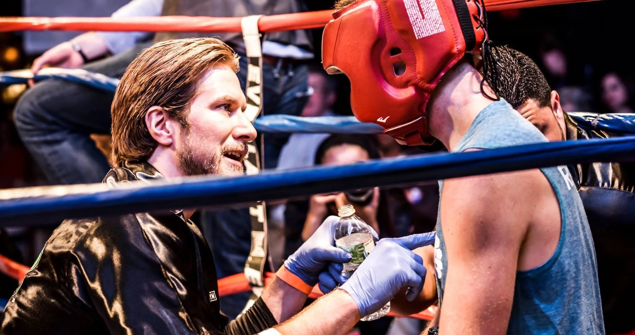 A boxing coach or trainer speaking intensely to a boxer in the ring, both wearing gloves, with the coach holding a water bottle and leaning in to communicate during a break in the match.