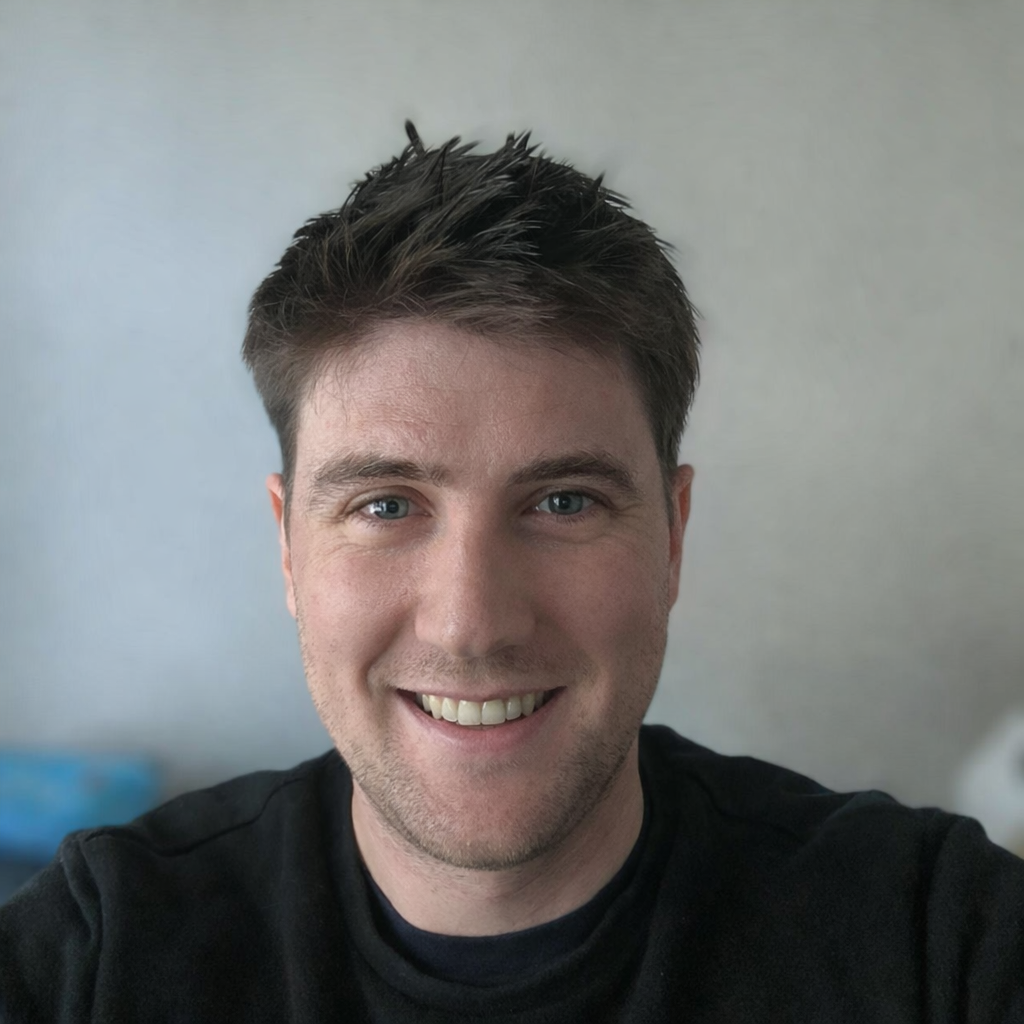 A young man with short, dark hair and blue eyes taking a selfie, smiling at the camera against a plain, light-colored background.