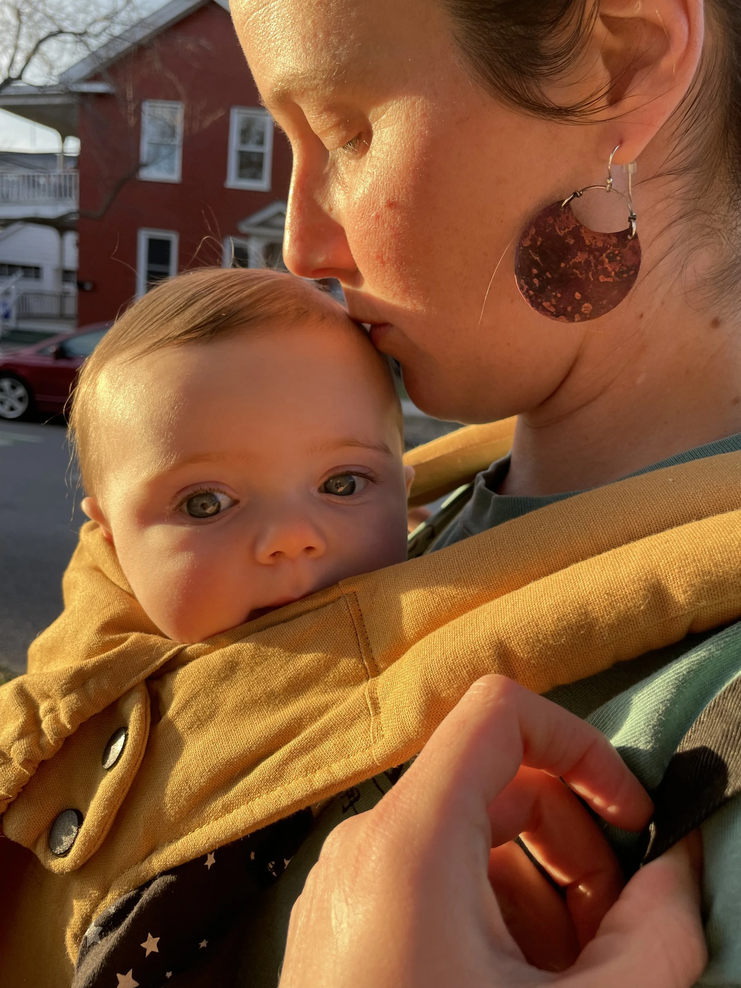 Doula, hypnobirthing and eft tapping practitioner, Lauren Akin holding her daughter in a yellow baby carrier while she kisses the top of her daughter's head during sunset.