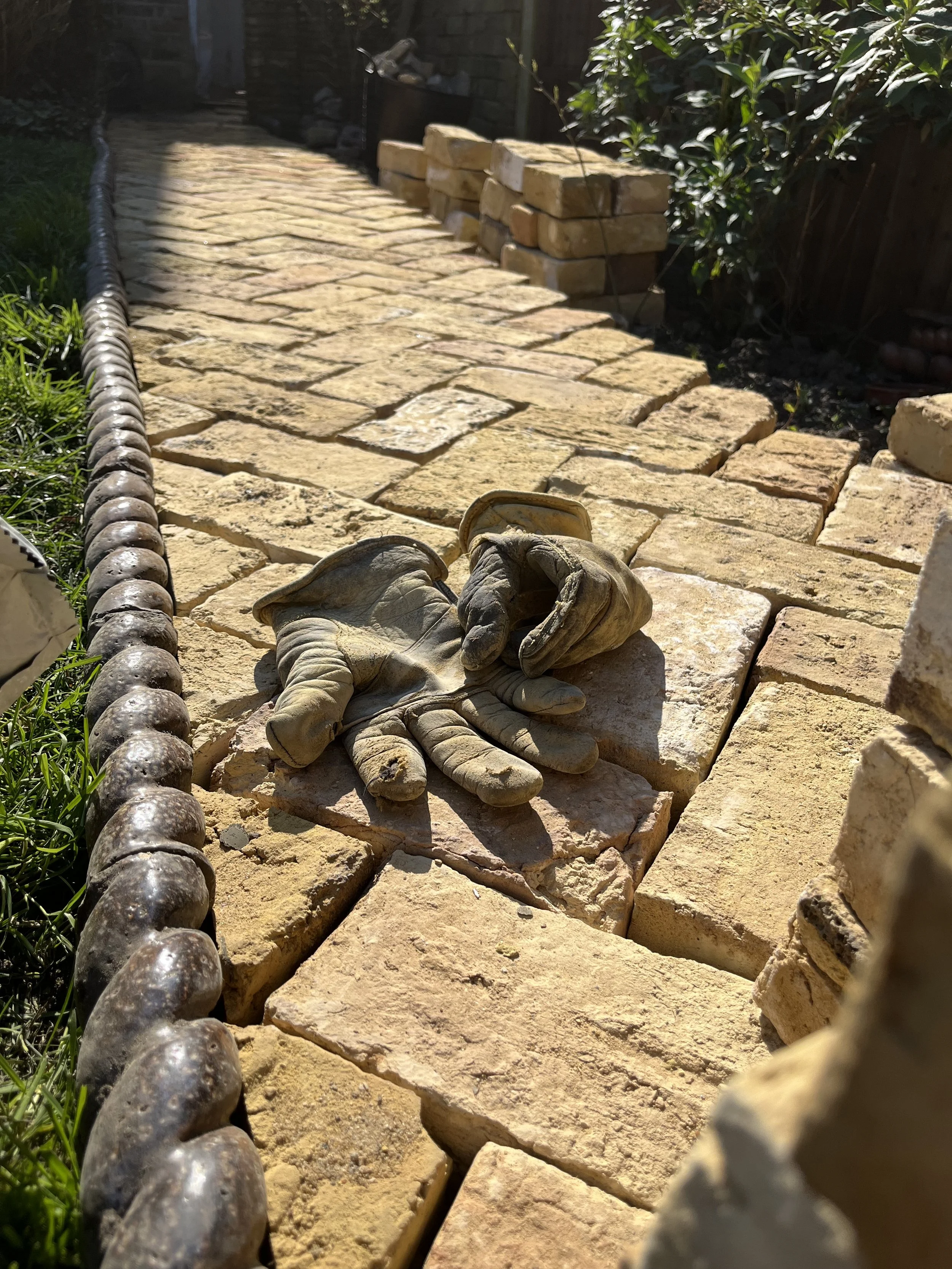 Close-up view of a stone pathway with a chain border on the left, a pair of work gloves placed on the stones, and some stacked bricks in the background. Sunlight casting shadows on the pathway. Green grass and plants along the edges.