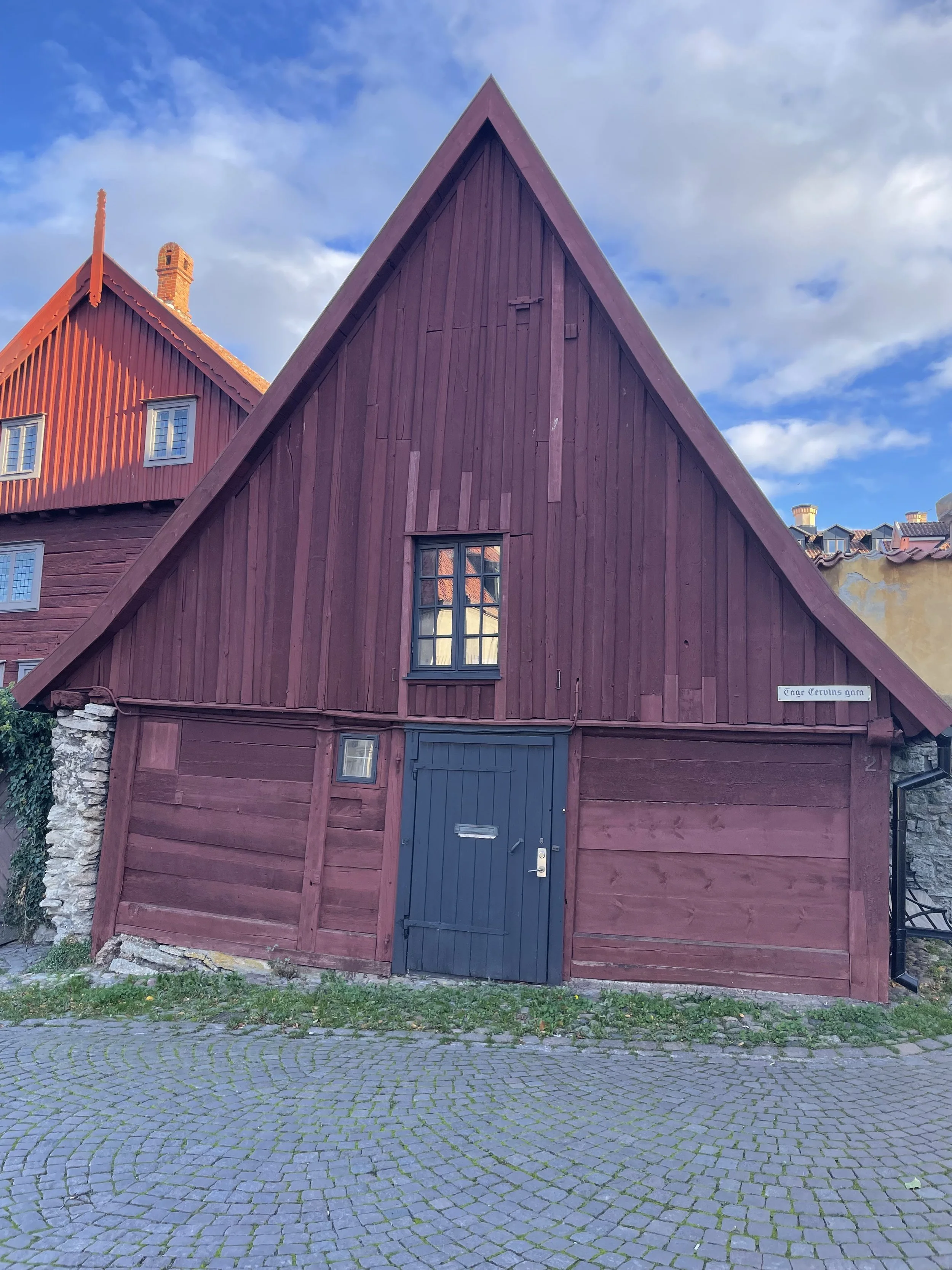 Red building with a steep roof, black door, and a small window, situated on cobblestone pavement with a cloudy sky in the background.